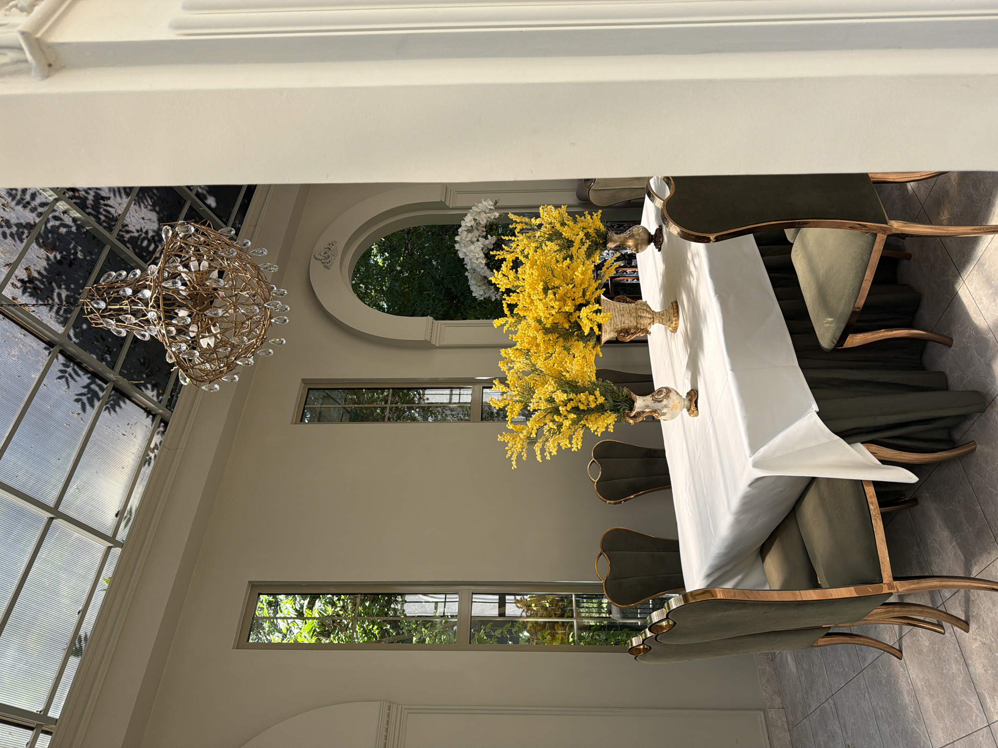 The image shows an indoor dining space, likely in Bishkek City, Kyrgyzstan. A long table covered with a white tablecloth dominates the scene, adorned with two vases filled with yellow flowers. Chairs with olive green upholstery and ornate wood frames surround the table. A decorative chandelier hangs from the ceiling. Natural light streams in through large windows and an arched opening, revealing trees outside.