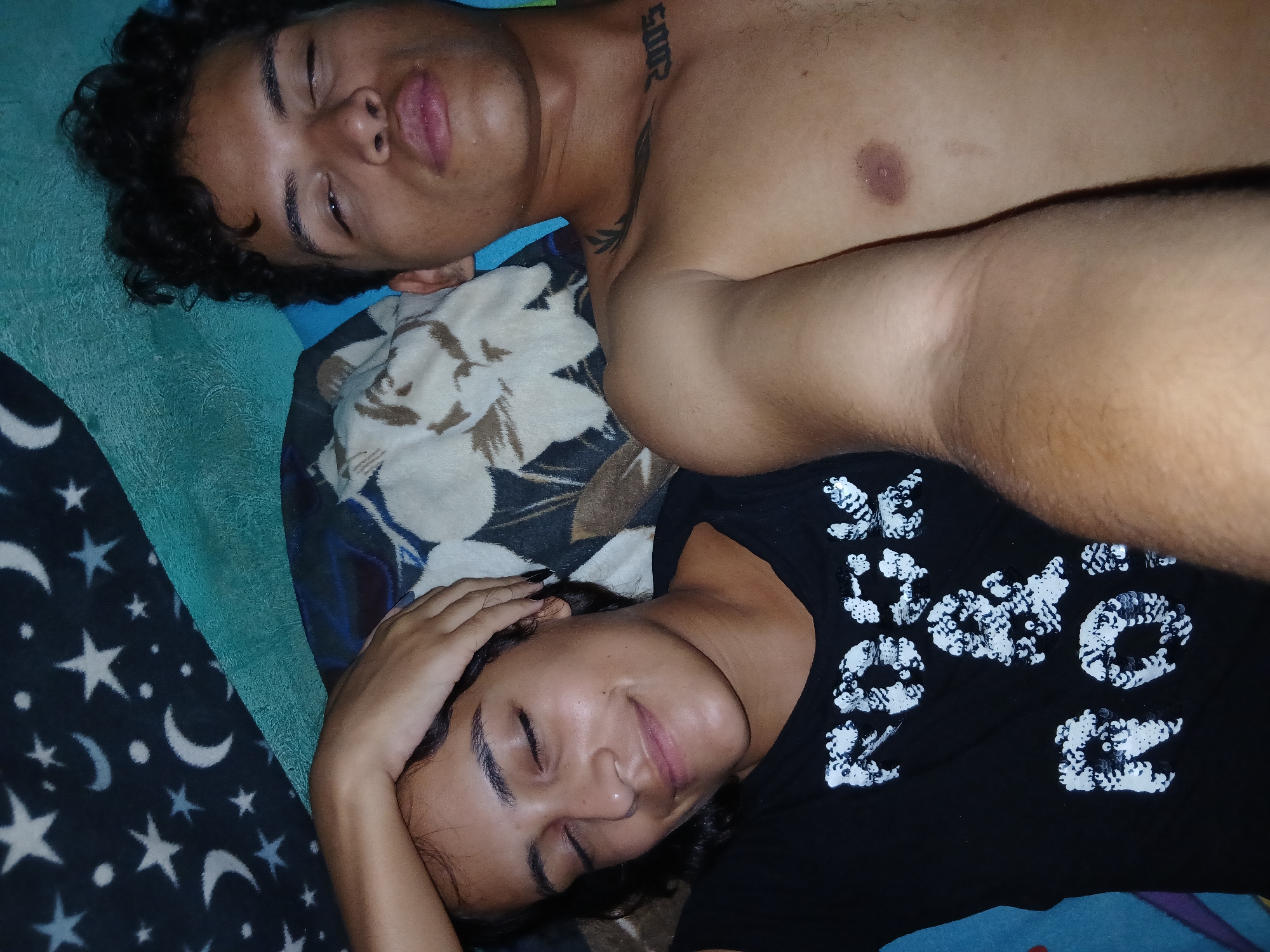 Two people are lying down on a bed. The person on top is a shirtless male with curly hair and a tattoo on his upper chest. The person below is wearing a black t-shirt with white sequin designs and has one hand on their forehead. They are both lying on a patterned pillow. The background is a blue blanket with a star and moon pattern. The location is not specified in the image.