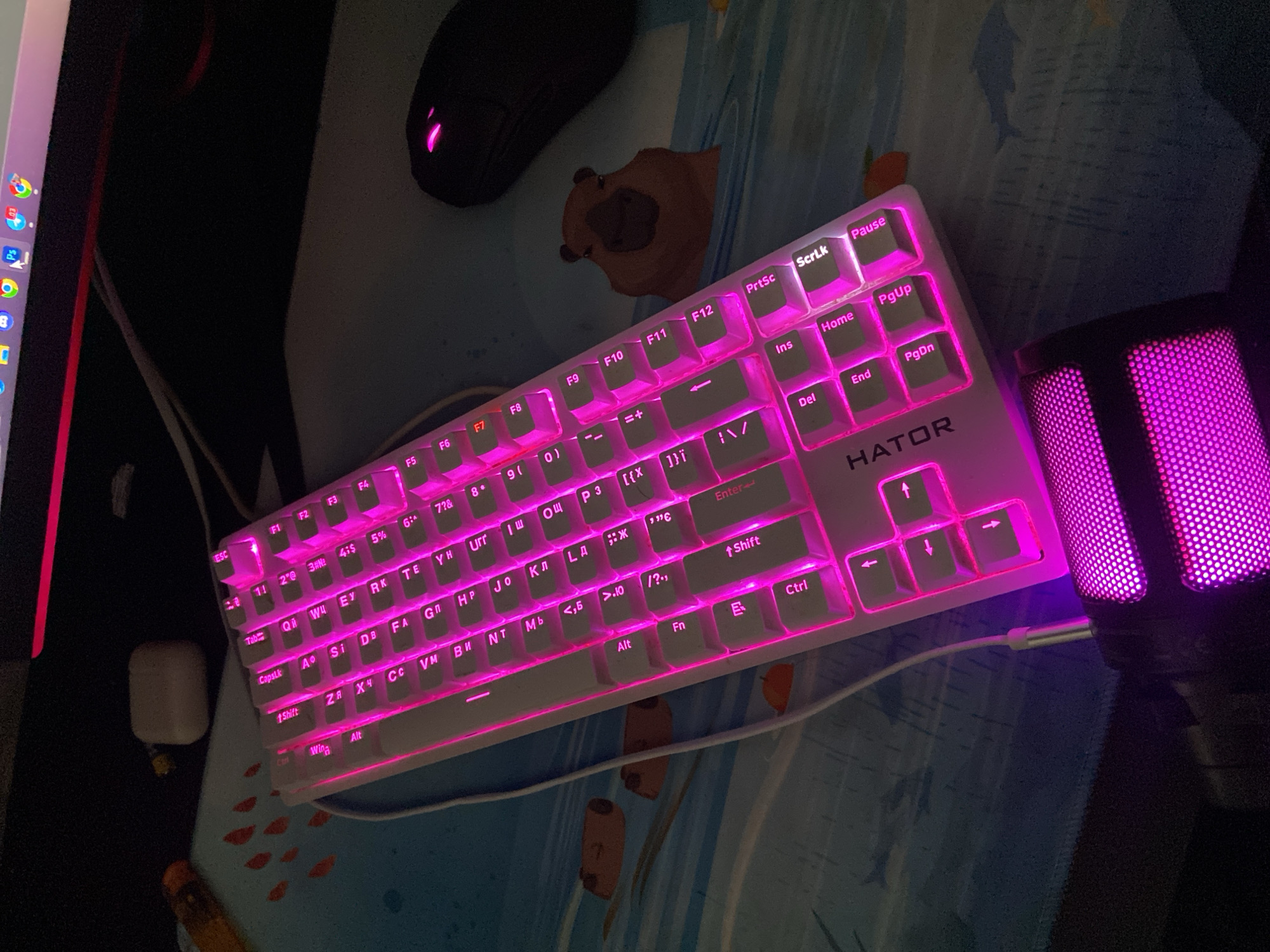 This is an image of a desk setup with a computer keyboard, mouse, and microphone. The keyboard is white with pink backlighting and has the brand name "HATOR" visible. The keys display a mix of English letters and Cyrillic characters, suggesting it may be a bilingual keyboard. The mouse, also black, has pink glowing accents. A pink-lit microphone stands to the right of the keyboard. The scene is illuminated by the pink glow of the peripherals, creating a vibrant, gaming-oriented atmosphere. The background shows a desk mat with a blue and white striped pattern and some small red fish designs. The time of day appears to be night or in a dimly lit room, as indicated by the dark surroundings and the bright backlighting of the keyboard and microphone. The overall impression is that of a functional and aesthetically pleasing setup for computer use, possibly for gaming or streaming. Visible text includes key labels on the keyboard such as "ESC", "F1" through "F12", "Shift", "Ctrl", "Alt", "Fn", and "Enter", as well as the brand name "HATOR". There are also Cyrillic characters present on many of the keys, such as "ОЩ" and "УН".
