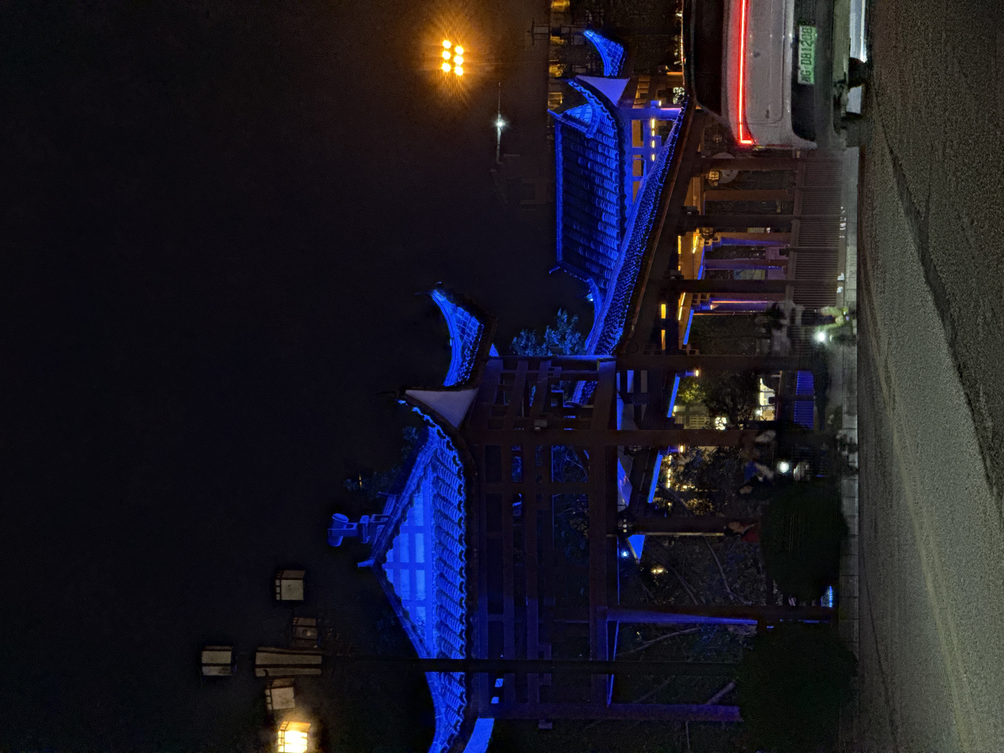 The image depicts a night scene in Wulingyuan, China, featuring traditional Chinese architecture illuminated with blue lights. The buildings appear to be pavilions or gazebos, likely part of a park or tourist area. The scene includes bodies of water, with lanterns or lights reflected on the surface. The presence of streetlights and what appears to be a parked car indicates an urban environment. The focus is on the architecture and lighting, creating a vibrant and atmospheric scene.