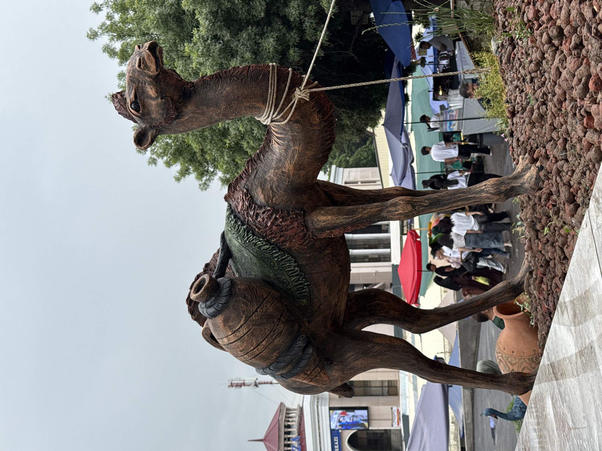 The image depicts a sculpture of a camel in a public setting. The camel is brown and appears to be made of a composite material. It has rope around its neck and a pack on its back fashioned like a jar. The sculpture stands amidst a crowd of people under a canopy of umbrellas and awnings, suggesting an outdoor market or festival. A building with architectural features is visible in the background, potentially a landmark in the area. The ground around the sculpture is covered with small brown pebbles. The location is Tashkent, Uzbekistan, based on visual clues and context.