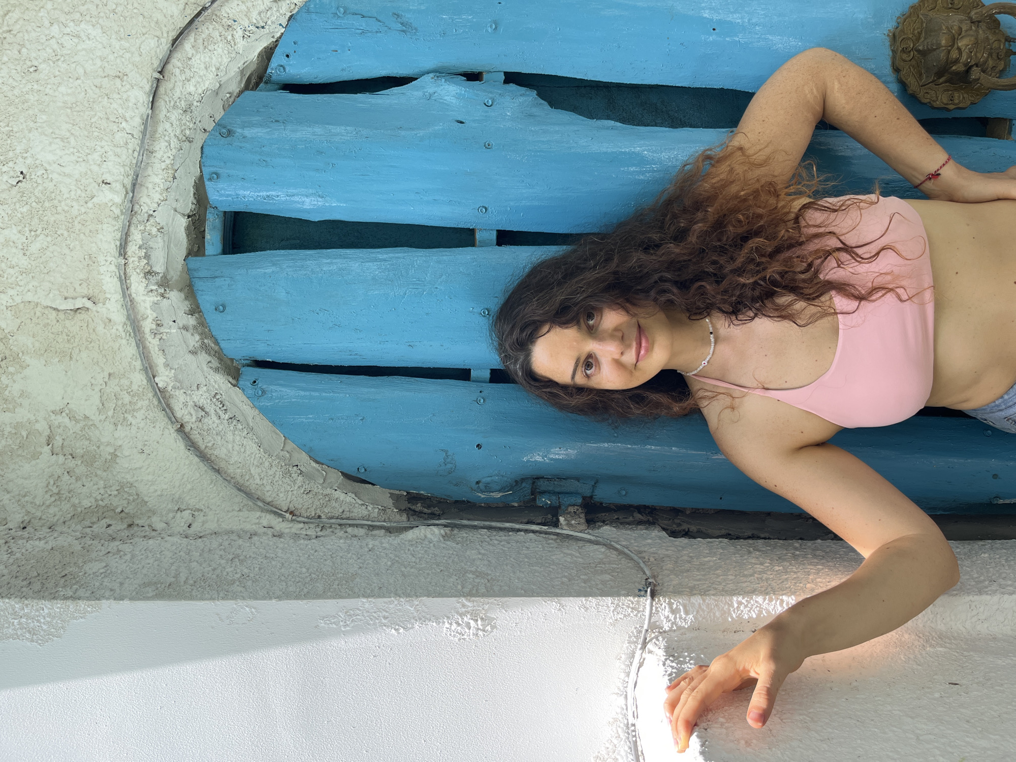 A woman with long, curly brown hair and a light complexion stands in front of a weathered, bright blue wooden door. She is wearing a pink halter-neck top and appears to be leaning against a white textured wall on the left side of the frame. Her left hand is placed on the wall, and her right arm is bent at the elbow with her hand resting on her hip. The door is composed of vertical wooden planks, some of which are split and uneven, with visible screws and hinges. Above the door, the wall is rough and textured, with an arched opening framed by weathered plaster. A metal wire runs along the wall and over the arch. To the right of the woman, a decorative lion head door knocker is attached to the blue door. The overall setting suggests an outdoor location, possibly in a historic or rustic part of a city. There is no visual information in the image to confirm the location as Phu Thuy Ward, Vietnam.
