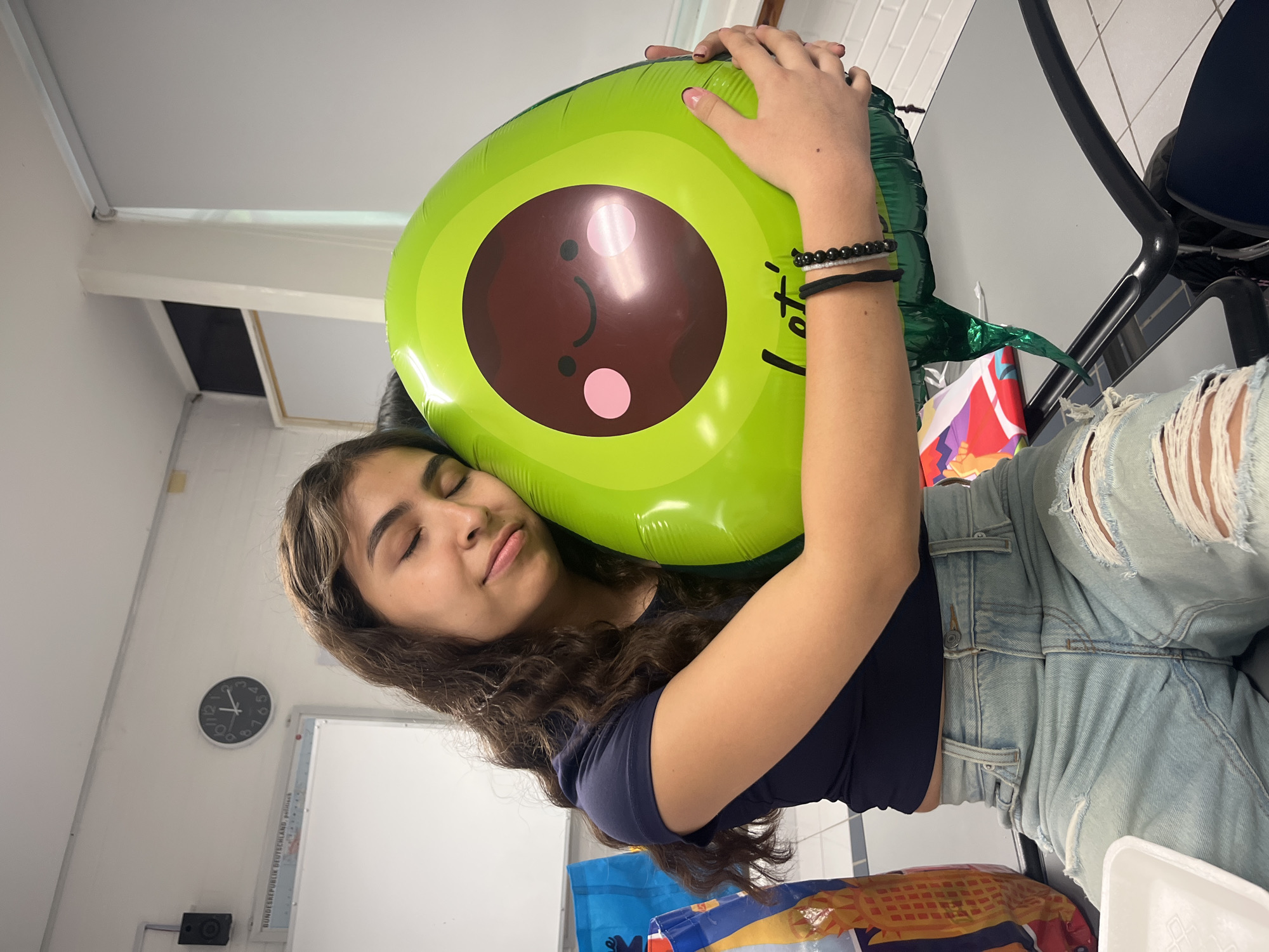 A young woman is shown holding an avocado-shaped balloon. She is wearing a dark-colored crop top and ripped light-wash denim jeans. Her eyes are closed and she appears to be relaxed or sleeping. The background includes a white wall with a clock and what appears to be a whiteboard. A table, chair, and other objects are also visible in the scene. The location is unknown.