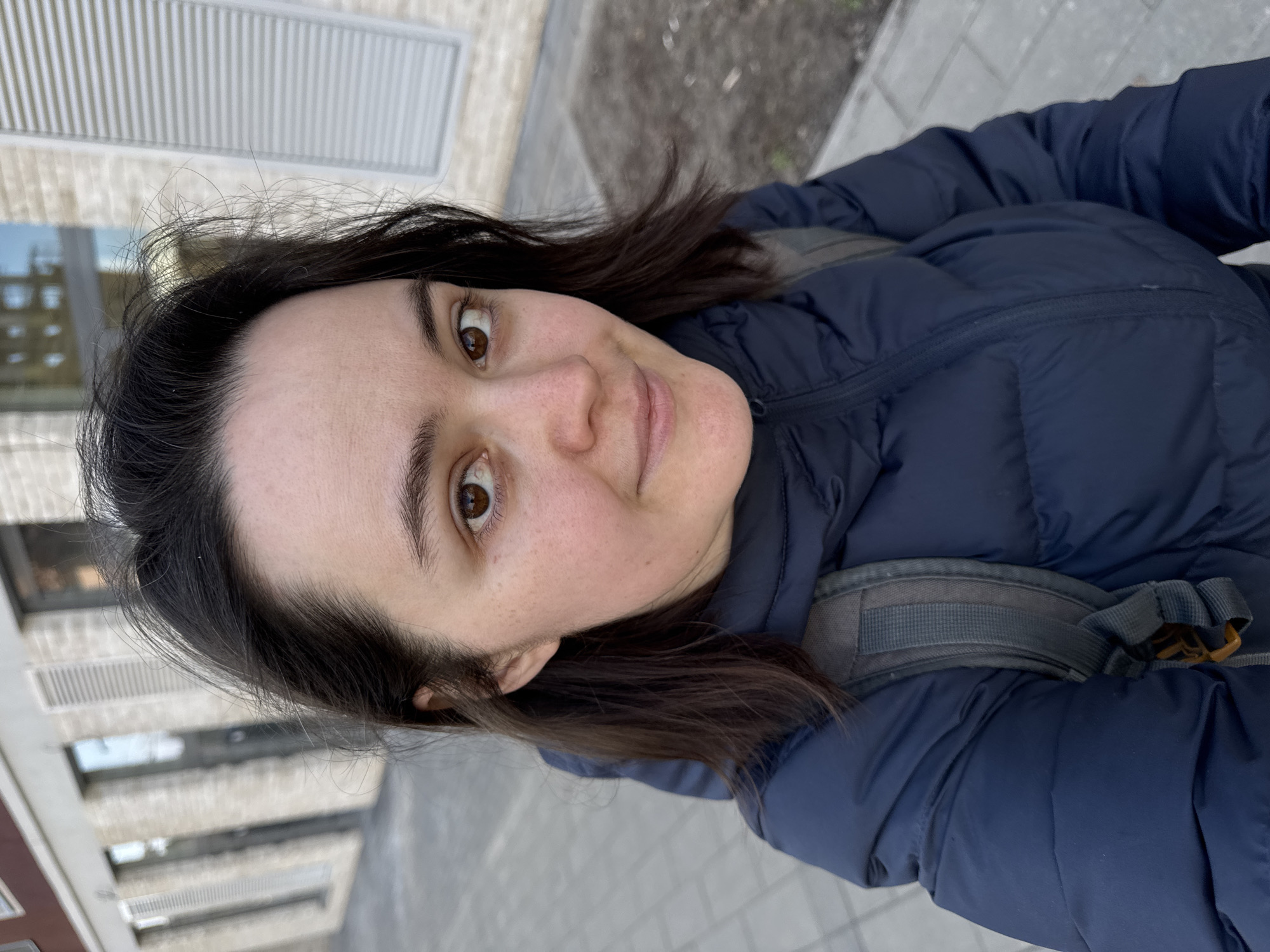 The image is a close-up, slightly tilted portrait of a woman outdoors in Izhevsk, Russia. She is looking directly at the camera with wide, brown eyes and a neutral expression that hints at a soft smile. Her dark brown hair is visible, appearing slightly windswept, and her complexion is fair with visible freckles on her nose and cheeks.
The woman is dressed for cool weather, wearing a dark blue puffer jacket. A grey backpack with visible straps and a buckle is worn over her right shoulder, indicating she might be traveling or commuting.
The background suggests an urban or suburban environment. Behind her, a light-colored building with prominent horizontal slatted vents or architectural features is visible. Other parts of the background show what appears to be another multi-story building with reflections in windows or structured balconies. The ground beneath her is paved, resembling a sidewalk or an urban pedestrian area.
The lighting suggests it is daytime, with soft, even illumination that could indicate an overcast sky or bright ambient light without direct sun. No visible text is present in the image. The woman appears to be taking a selfie or being photographed from a close distance while outdoors.