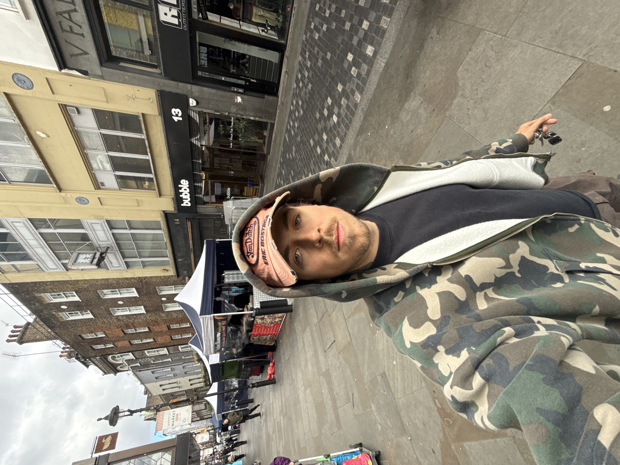 The image shows a man wearing a camouflage hoodie and a baseball cap standing on Berwick Street in London, United Kingdom. In the background, there are various buildings with shops, including "bubble" at number 13 and a market. The man is holding keys in his hand. The street is paved and appears to be busy with pedestrians.