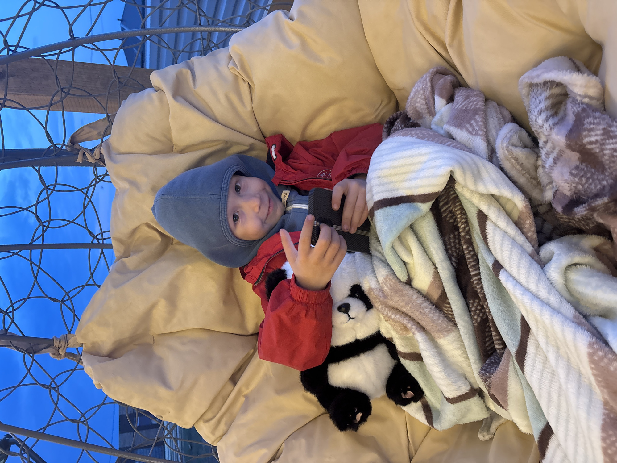 A young child, likely a toddler or preschooler, is lying in a cushioned hanging chair outdoors. The child is wearing a blue hooded hat, a red jacket, and what appears to be a grey-blue undershirt. The child is holding a black object in their hand and is also cuddling a plush panda bear. The chair is draped with a striped blanket and is suspended by a metallic frame. The background shows a blurred view of the sky and a building, suggesting the photo was taken outdoors during daylight. The location is Chaikovsky, Russia.