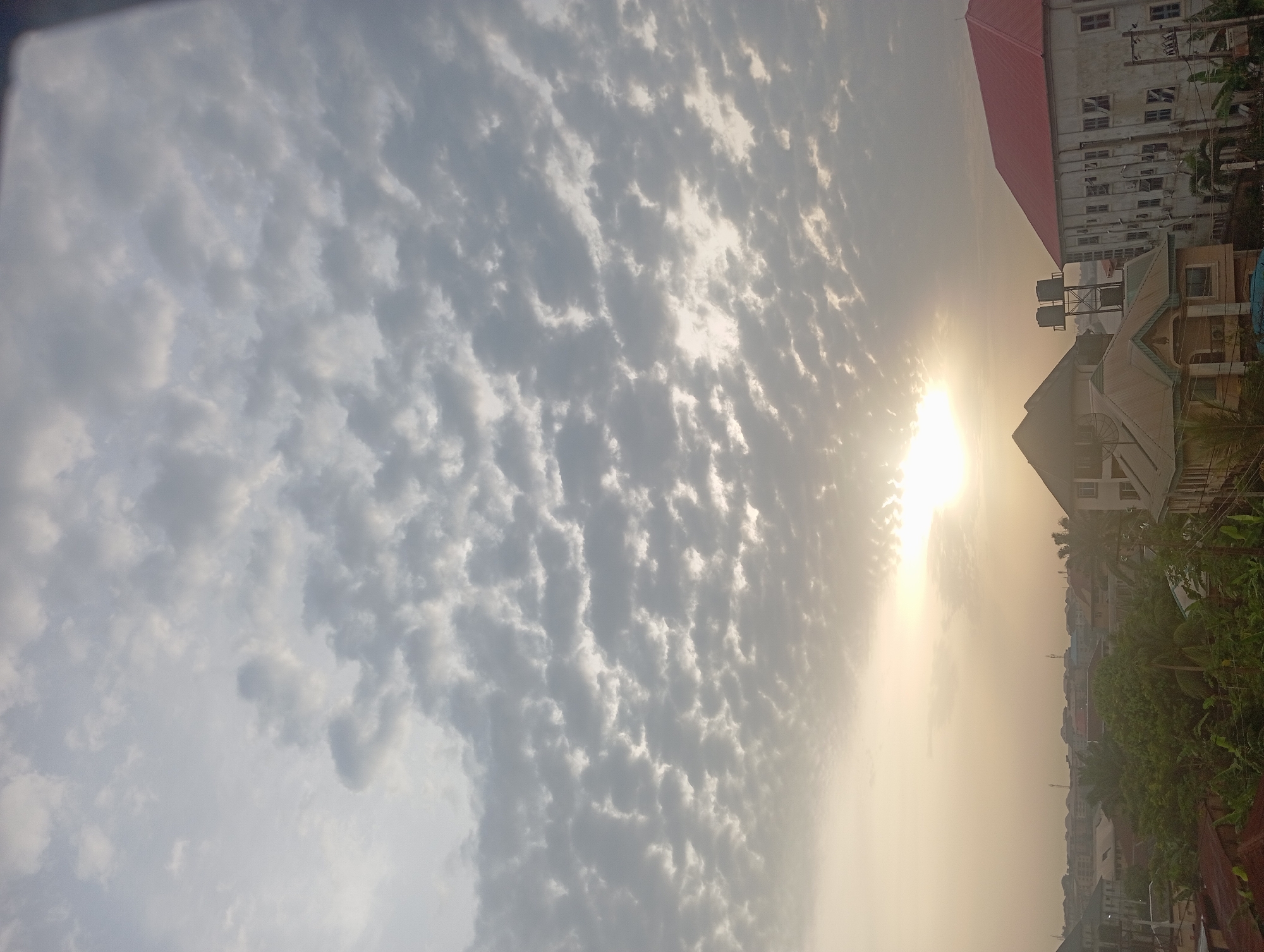The image shows a daytime scene in Nkpor, Nigeria. The sky is filled with altocumulus clouds, with the sun shining brightly through a gap in the cloud cover. The sun's rays create a hazy, golden light. Buildings and foliage are visible in the lower part of the image. Some buildings are tall with multiple windows and a red roof; other buildings have a beige, pitched roof. Green foliage is interspersed with the buildings. Water tanks are visible on the roofs of some buildings. No people are seen in the image.