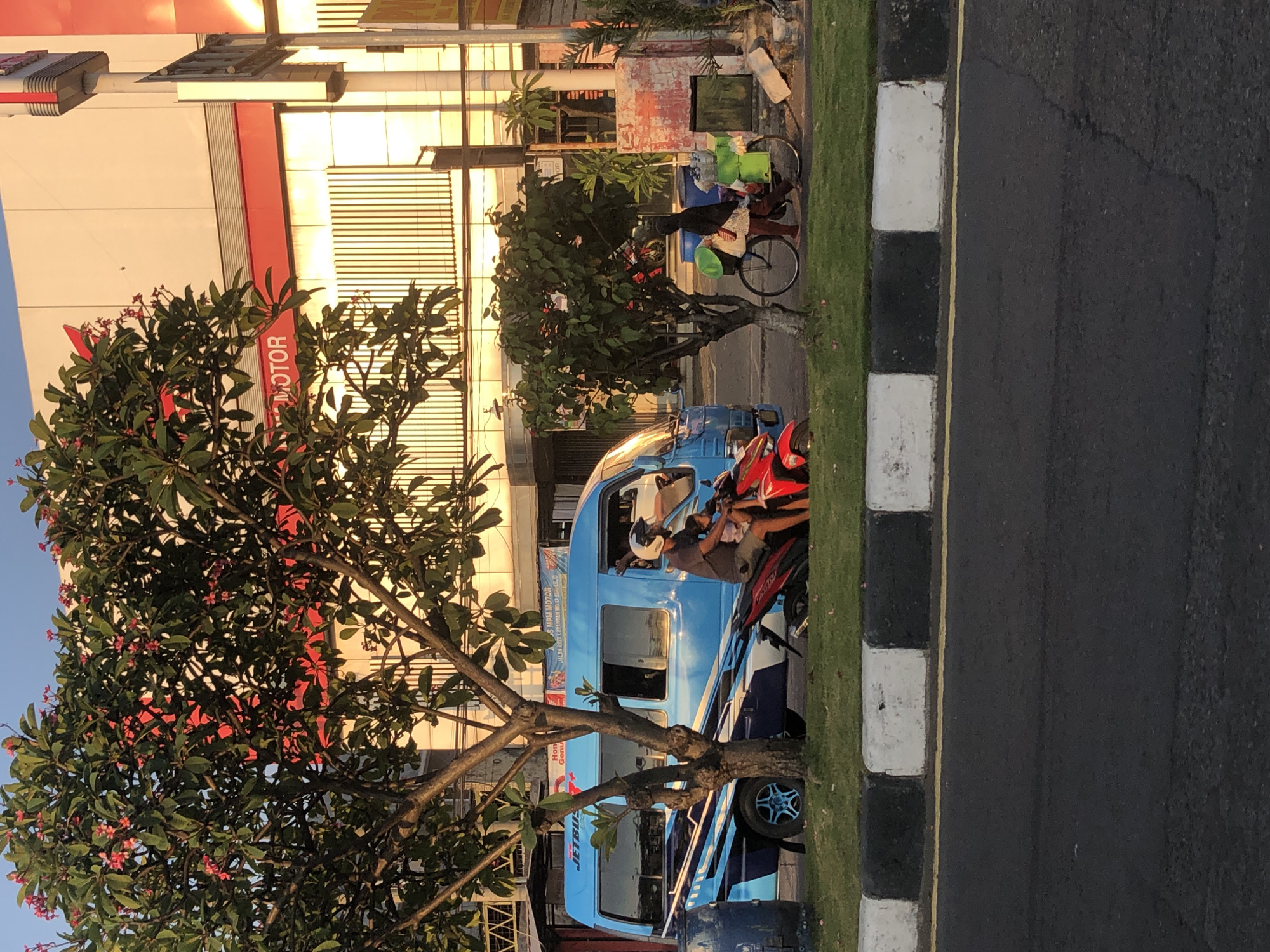 The image shows a street scene in Larangan, Indonesia, with a blue van and a red motorcycle parked on the side of the road. A man wearing a helmet is riding the motorcycle with a child sitting behind him. Behind them, a woman on a bicycle is pushing it with a cart attached to it, loaded with items. The background features buildings with a red and white facade, and trees with pink flowers. The street is paved, with a sidewalk demarcated by black and white concrete blocks. The scene appears to be captured during daylight.
