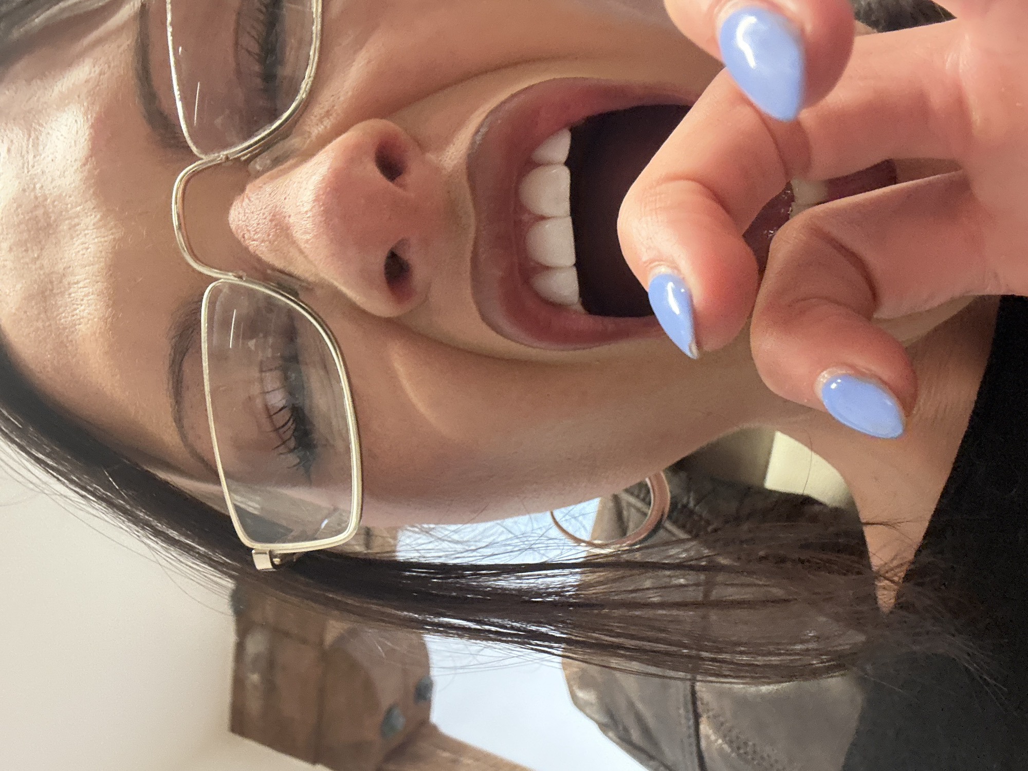 A close-up, vertically-oriented image depicts a woman with her head tilted downwards. She is wearing silver-rimmed glasses and has long, dark hair. Her mouth is open, revealing white teeth. Her fingers are near her mouth, adorned with light blue nail polish. The background is out of focus, showing what appears to be interior elements and clothing. The location is not identifiable from this image.