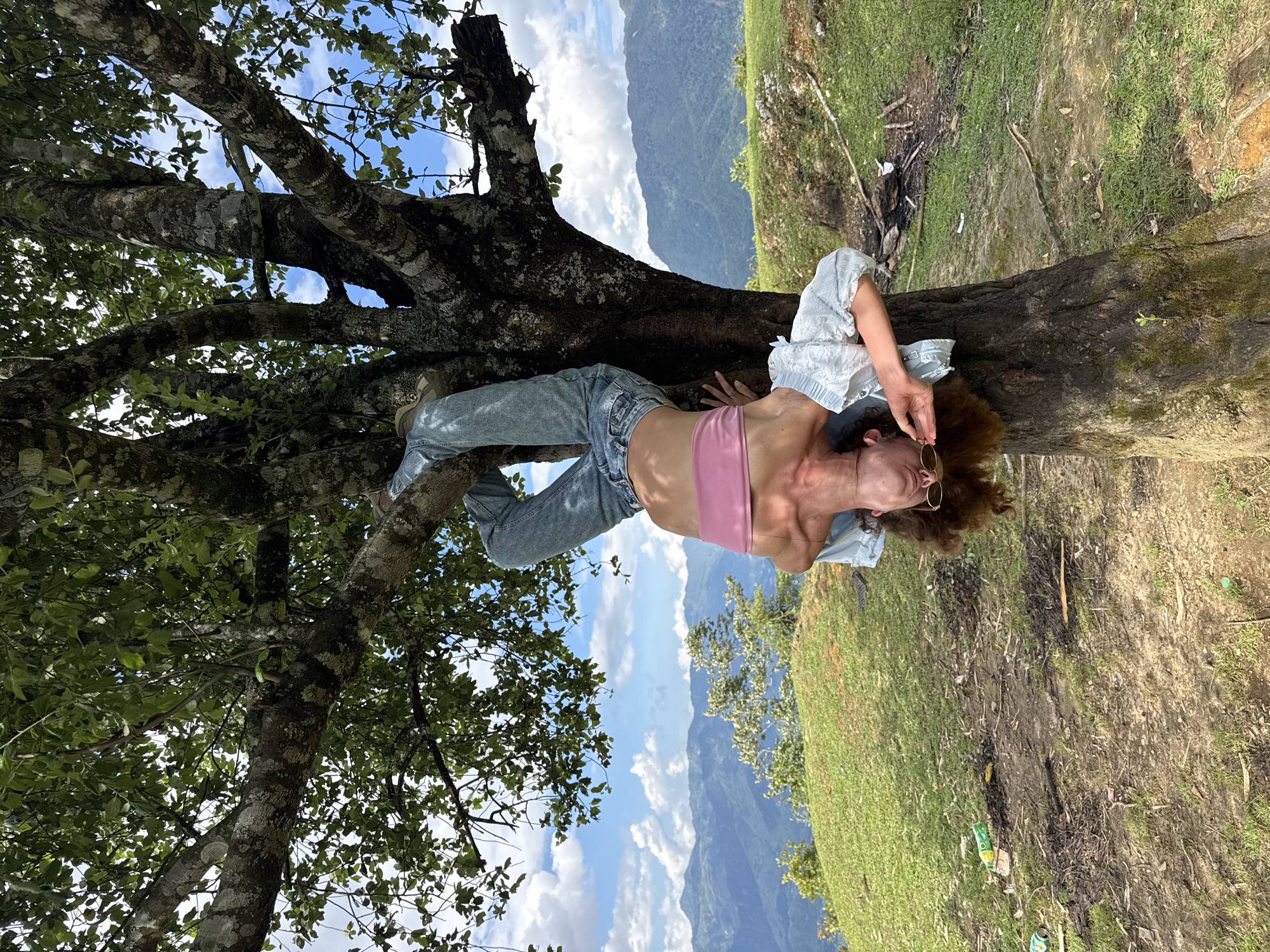 The image displays one individual hanging upside down from a large, mature tree in Tu Le, Vietnam. The person is suspended by their legs, which are wrapped around a substantial tree branch. They are attired in light blue denim jeans, a pink strapless top, and a light-colored, short-sleeved patterned blouse worn over the pink top. Their long, curly brown hair hangs freely towards the ground, and small, round-framed sunglasses cover their eyes, with one hand positioned near the face. The tree features a thick trunk and branches with textured, mottled grey and brown bark, supporting a dense canopy of green foliage. In the background, a multi-layered mountainous landscape stretches beneath a blue sky dotted with white clouds. The foreground shows a gently sloping terrain composed of green grass interspersed with patches of bare earth, containing scattered human-made debris such as plastic bottles and aluminum cans, alongside natural litter and evidence of burnt material.