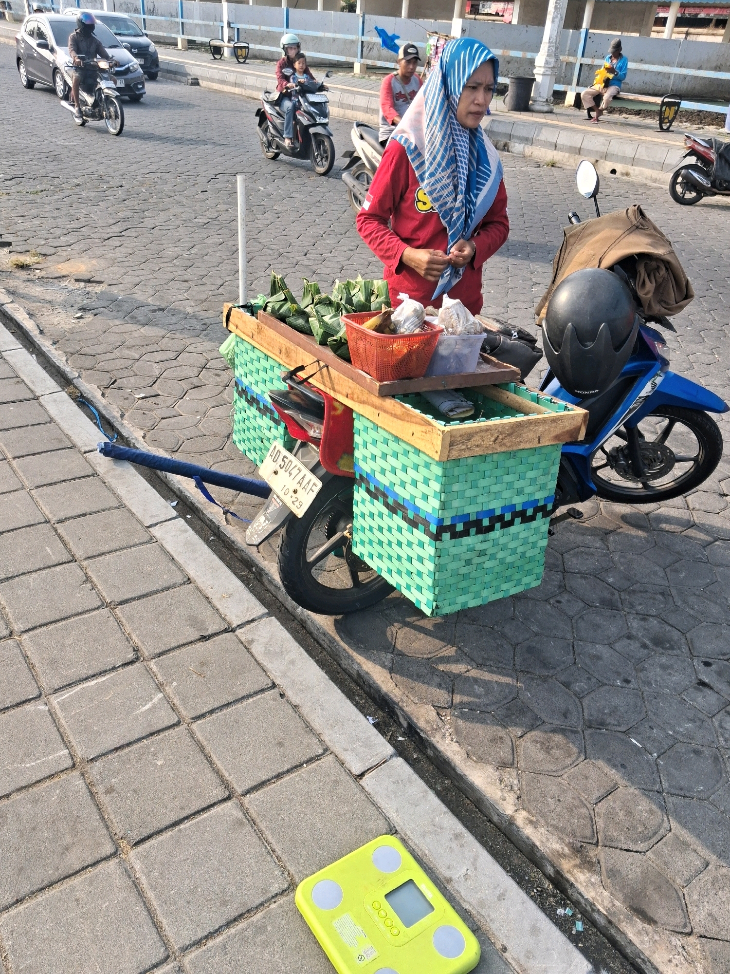 In the city of Surakarta, Indonesia, a woman wearing a blue and white hijab and a red long-sleeved shirt stands beside a motorcycle fitted with a green woven basket cart. She is holding a small item in her hands and appears to be engaged in a transaction. The cart contains packaged food items, a red basket with bananas and other packaged goods, and a black helmet. The motorcycle is blue and red, with a license plate reading "AD 5047 AAF". In the foreground, a yellow digital scale rests on a paved sidewalk. The background shows a street with several other motorcycles and cars, as well as a bridge and a raised sidewalk with people seated on a bench. The scene is set on a cobblestone street with a drainage ditch along the side.