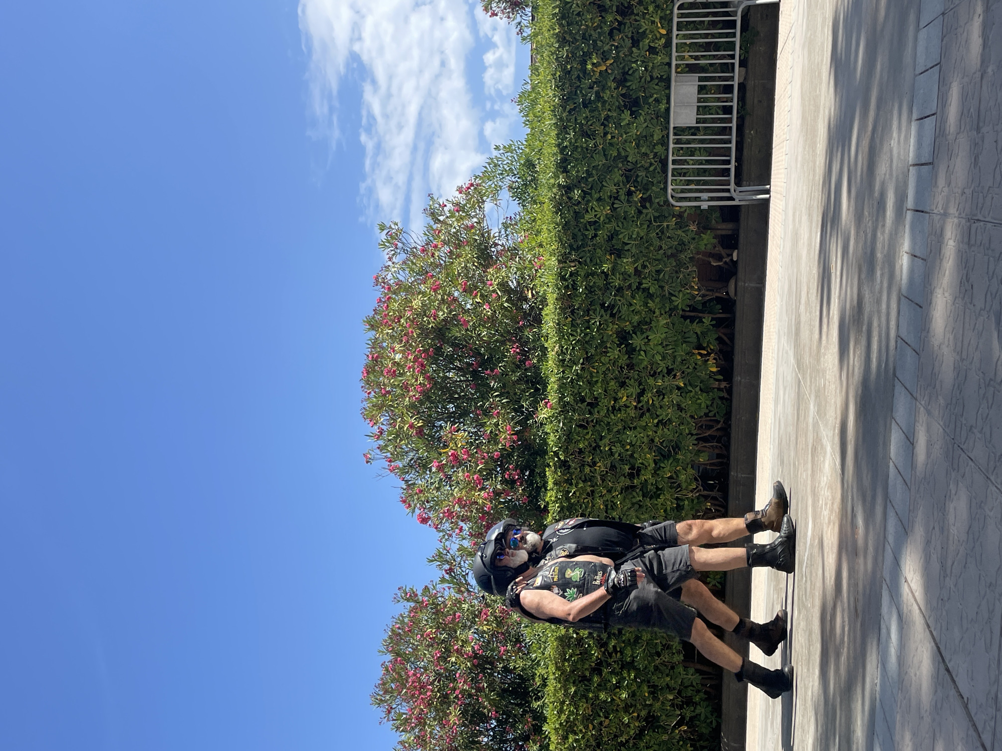 Two men walk side-by-side along a paved walkway in Sitges, Spain, with a meticulously trimmed green hedge and flowering pink bushes behind them. Both men are wearing black helmets and appear to be of similar build, with beards and sunglasses. The man on the left wears a black leather vest with patches over a black shirt, gray shorts, and black boots. The man on the right wears a black vest over a black shirt, black shorts, and brown boots. A metal crowd barrier is visible to the right of the hedge. The sky is a clear, bright blue with a few wispy clouds.