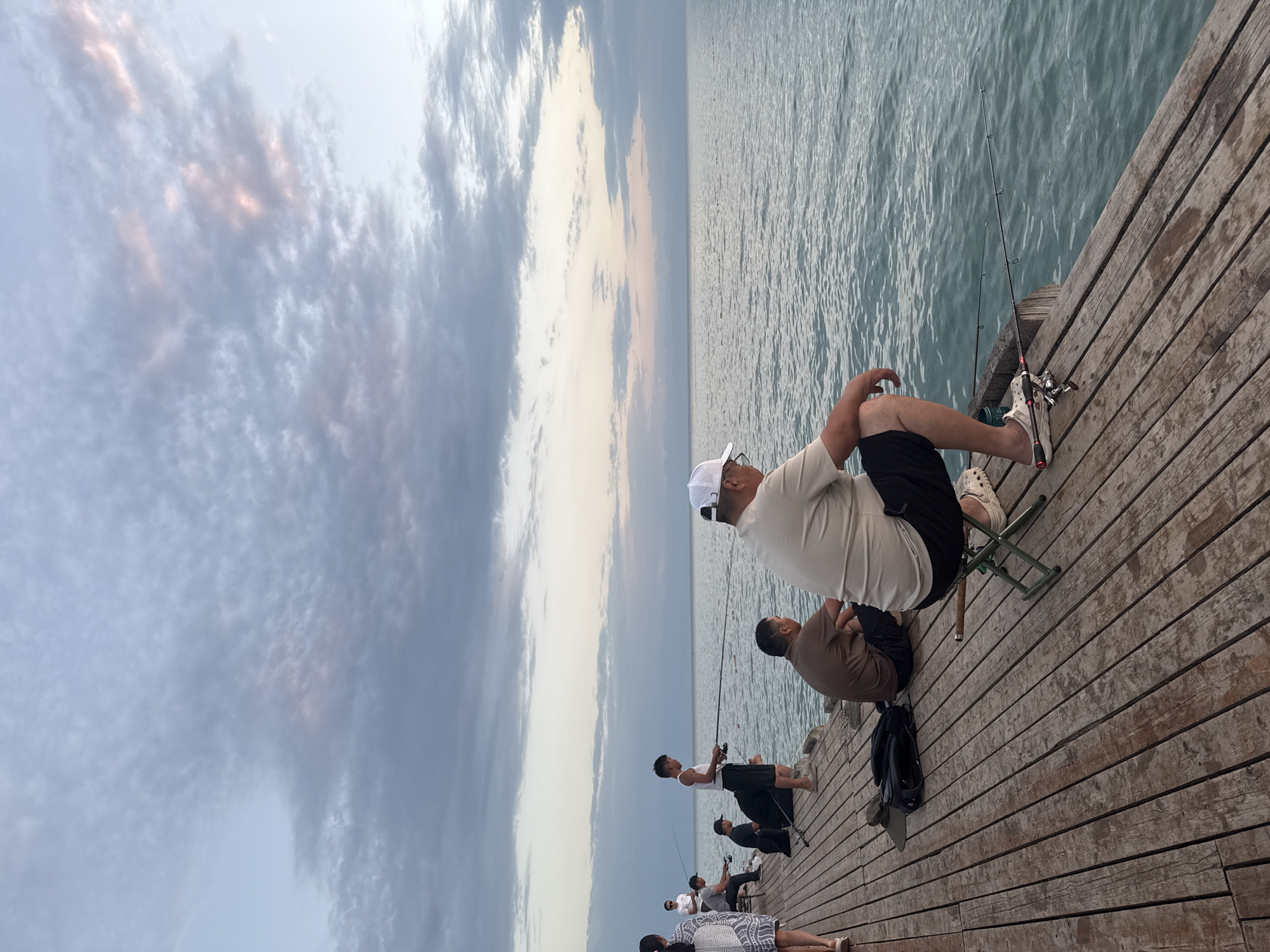A group of people are fishing from a wooden pier on the shore of Issyk-Kul Lake, in the city of Sary-Oy, Kyrgyzstan. The sky is filled with dramatic clouds, and the sun is setting, casting a warm glow on the scene. The water is calm, with gentle waves lapping against the pier. Several people are actively fishing, while others are watching the water or enjoying the view. The pier is made of weathered wood, and there are fishing rods and other equipment scattered around. The overall atmosphere is peaceful and serene, with a sense of community among the anglers.