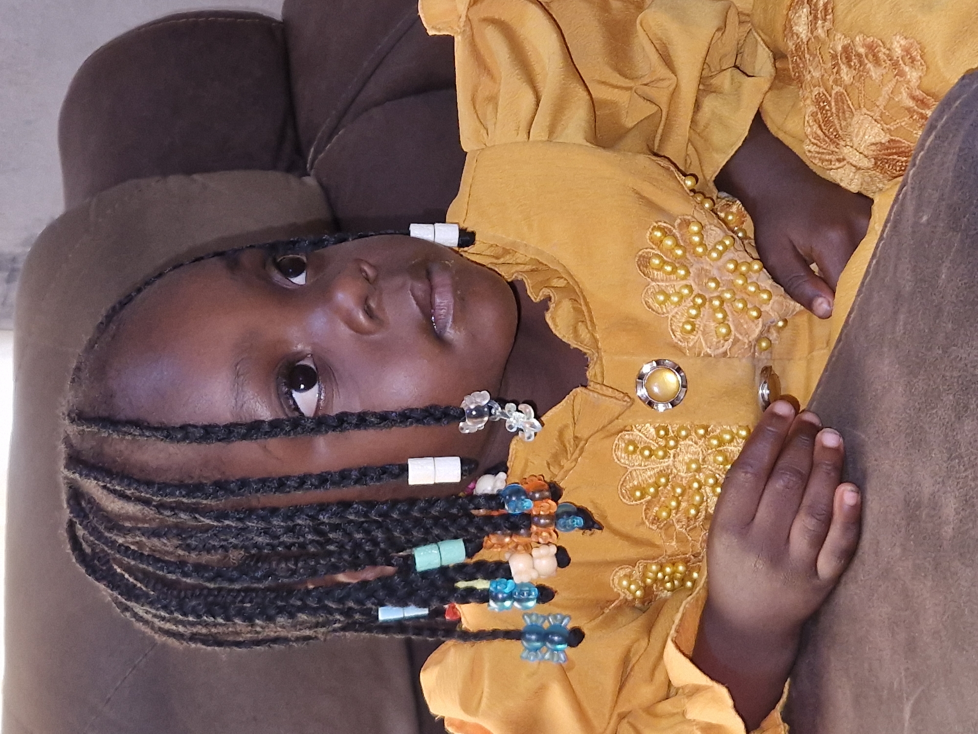 A young Black child, likely female due to the braided hair adorned with colorful beads and charms, is positioned in a medium shot, facing to the right. The child has dark skin and is wearing a bright yellow, ruffled dress with beaded embellishments. The child's left hand rests on her lap, and her right hand is also visible. She is seated on a brown upholstered surface, which appears to be a couch or chair. The background is a blurred brown. The scene appears to be indoors, possibly in a residential setting in Jos, Nigeria.