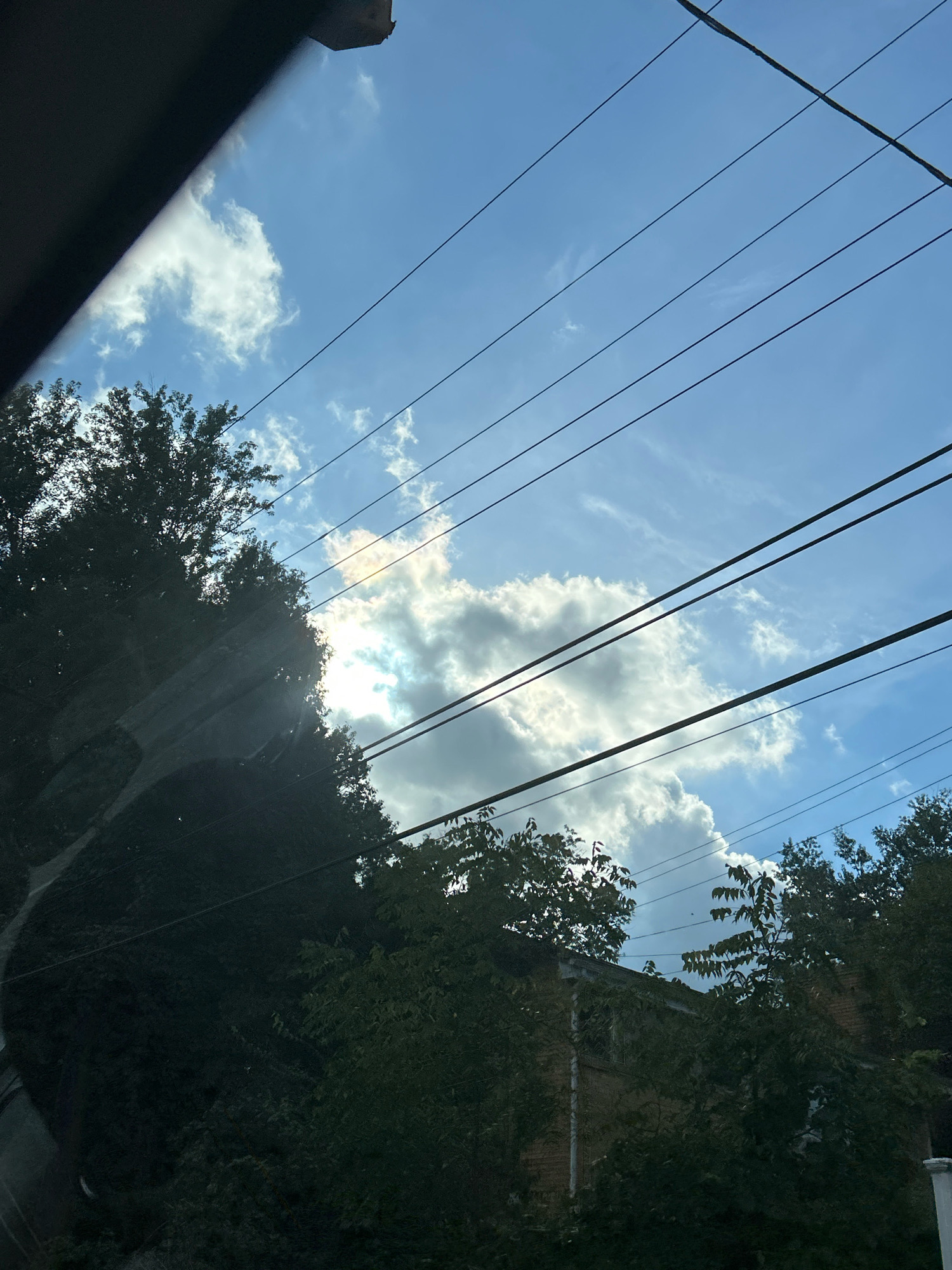 The image displays a view of the sky from a low-angle perspective, likely from a vehicle, looking upwards. Overhead power lines are prominently featured, stretching diagonally across the frame. The sky is blue with scattered white clouds. Sunlight breaks through a large cloud formation, creating a bright, luminous area with visible rays. Lush green trees partially obscure the lower portion of the scene, with some foliage appearing in silhouette against the bright sky. A portion of a building, constructed with brick, is visible beneath the trees. The overall impression is one of an outdoor, possibly residential, setting on a partly cloudy day, with elements of urban infrastructure like power lines and buildings interspersed with natural elements like trees and clouds. The location is not specified beyond the United States of America.
