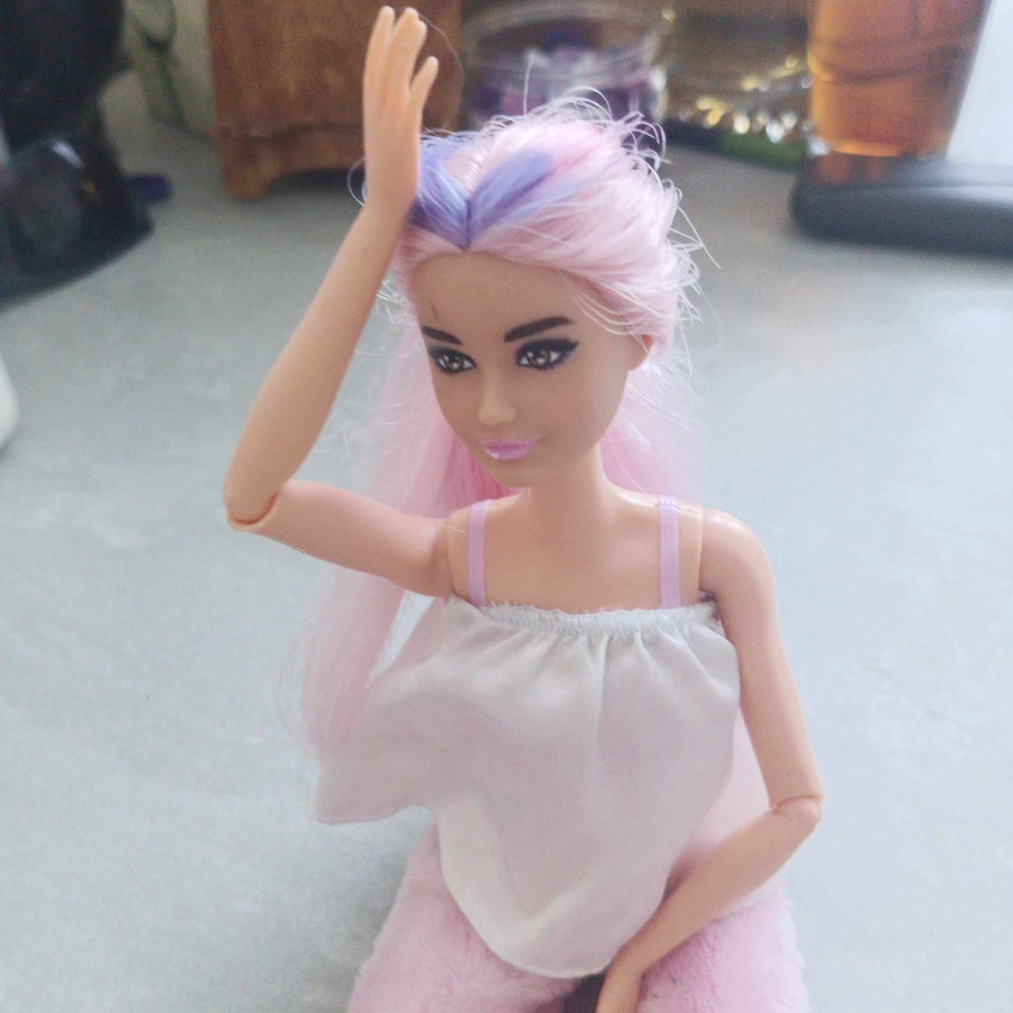 A Barbie doll with pink and purple hair, wearing a white top with pink straps and pink furry pants, is posed on a light gray surface. The doll's right arm is raised. In the blurred background, there are various objects including a black appliance, a wooden box, a jar, and a cup. The location and specific details about the city of Chaikovsky, Russia, cannot be determined from the given image.