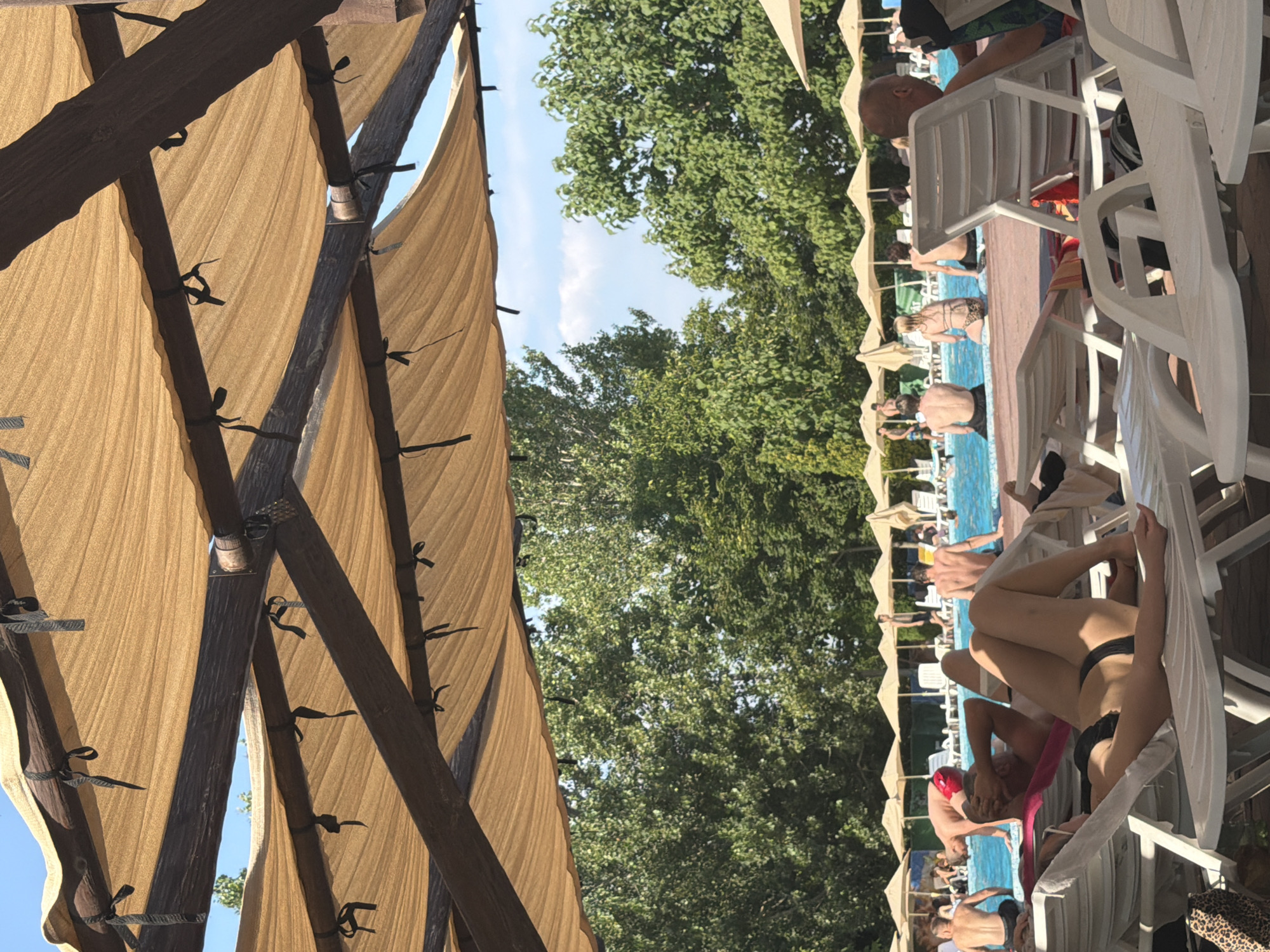 The image depicts a sunny day at a resort pool, possibly in Sociteni, Moldova, with numerous people enjoying the water and lounging on chairs under a large, fabric canopy. In the foreground, a woman in a black bikini reclines on a white plastic lounge chair, her legs extended. Behind her, a man also reclines on a chair, facing away from the camera, partially obscured by her. Other people are visible in the background, some sitting by the pool's edge, others wading or swimming in the blue water. Many white lounge chairs and beige umbrellas are arranged around the pool. Dense green trees form a backdrop to the scene, and the sky is a clear blue with a few wispy clouds. The canopy overhead, made of tan fabric stretched over a wooden frame, provides shade.