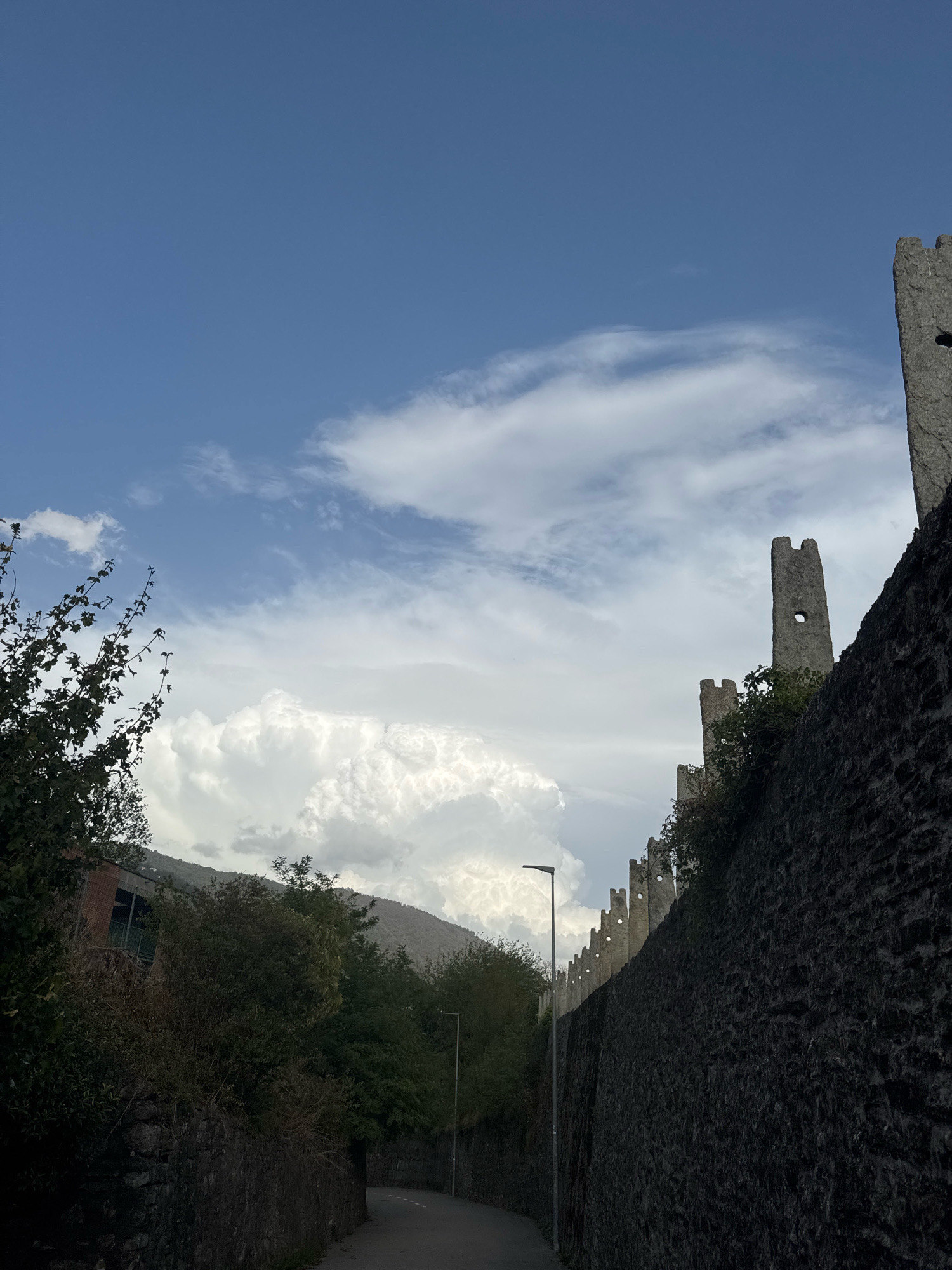 A steep, upward-angled shot captures a cobblestone path curving around a massive stone wall, part of a historic fortification. The wall is dark and textured, with a series of crenellated battlements and watchtowers rising along its edge. The sky above is a vibrant blue, punctuated by dramatic white clouds, some of which have a cumulonimbus appearance, suggesting recent or ongoing weather activity. Green foliage, including trees and bushes, spills over the wall in places and lines the path. To the left, a building with a red brick facade and large windows is visible amidst the trees. The overall scene suggests a historic and somewhat imposing location, possibly a castle or an ancient city wall, situated in a hilly or mountainous region given the backdrop of a distant, tree-covered mountain slope. The location is identified as Balerna, Switzerland, implying a European setting with a rich history.