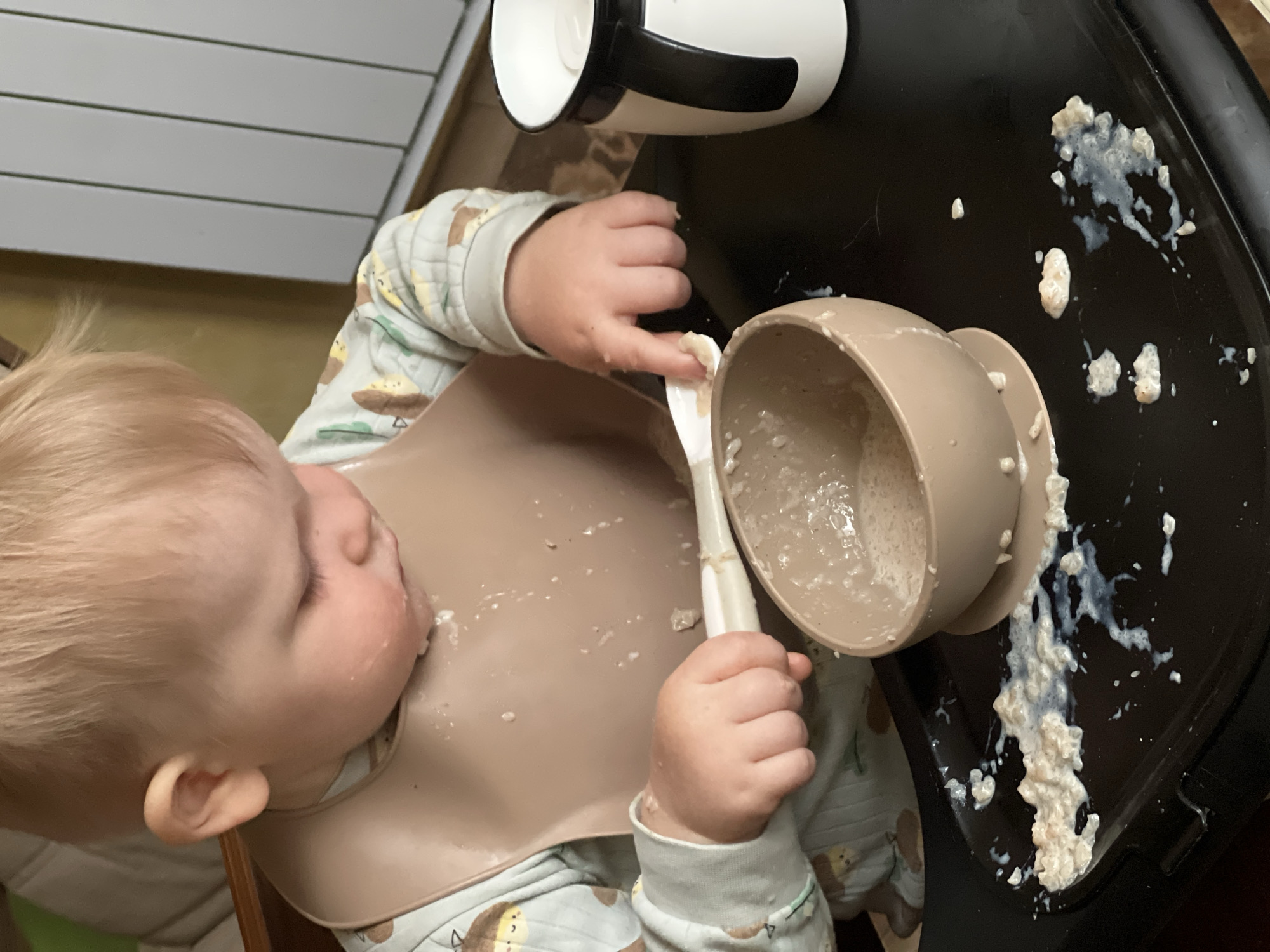 A young child with blonde hair is sitting at a highchair, wearing a light-colored pajama set with a dog or monkey pattern and a light brown bib. The child is holding a spoon in their right hand and appears to be eating from a light brown bowl. A white and black cup is visible on the table next to the bowl. The black tray of the highchair has splattered oatmeal or porridge on it. The image provides no information about Khimki, Russia.
