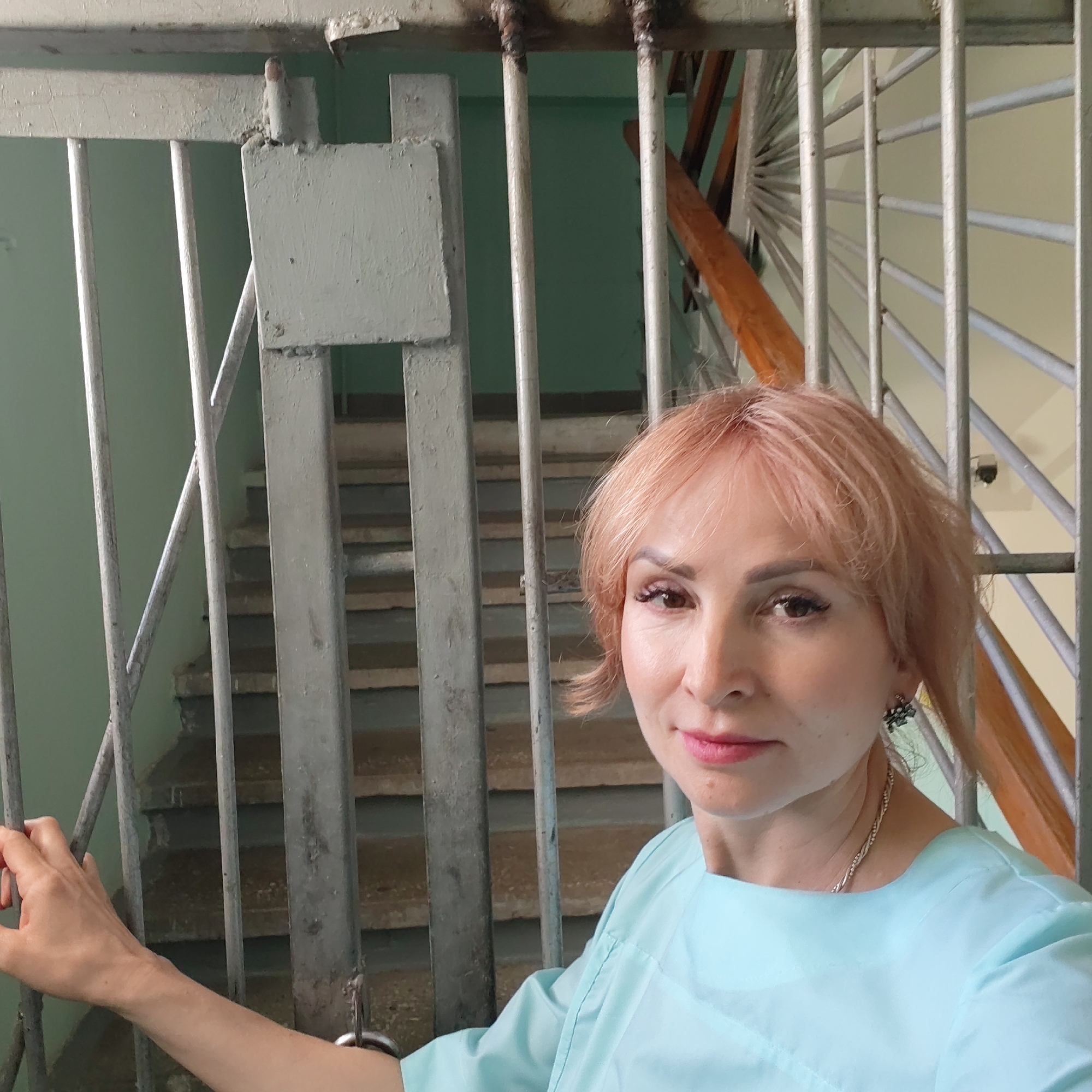 A woman with short, light pink hair stands in front of a metal security gate, likely in an urban setting in the city of Unknown, Unknown. She wears a light blue top, a silver necklace, and earrings with dark green accents. Her left hand rests on the gate. The gate appears to be a security measure for a stairwell visible in the background, with concrete steps and a metal railing. The wall is painted a light green color.