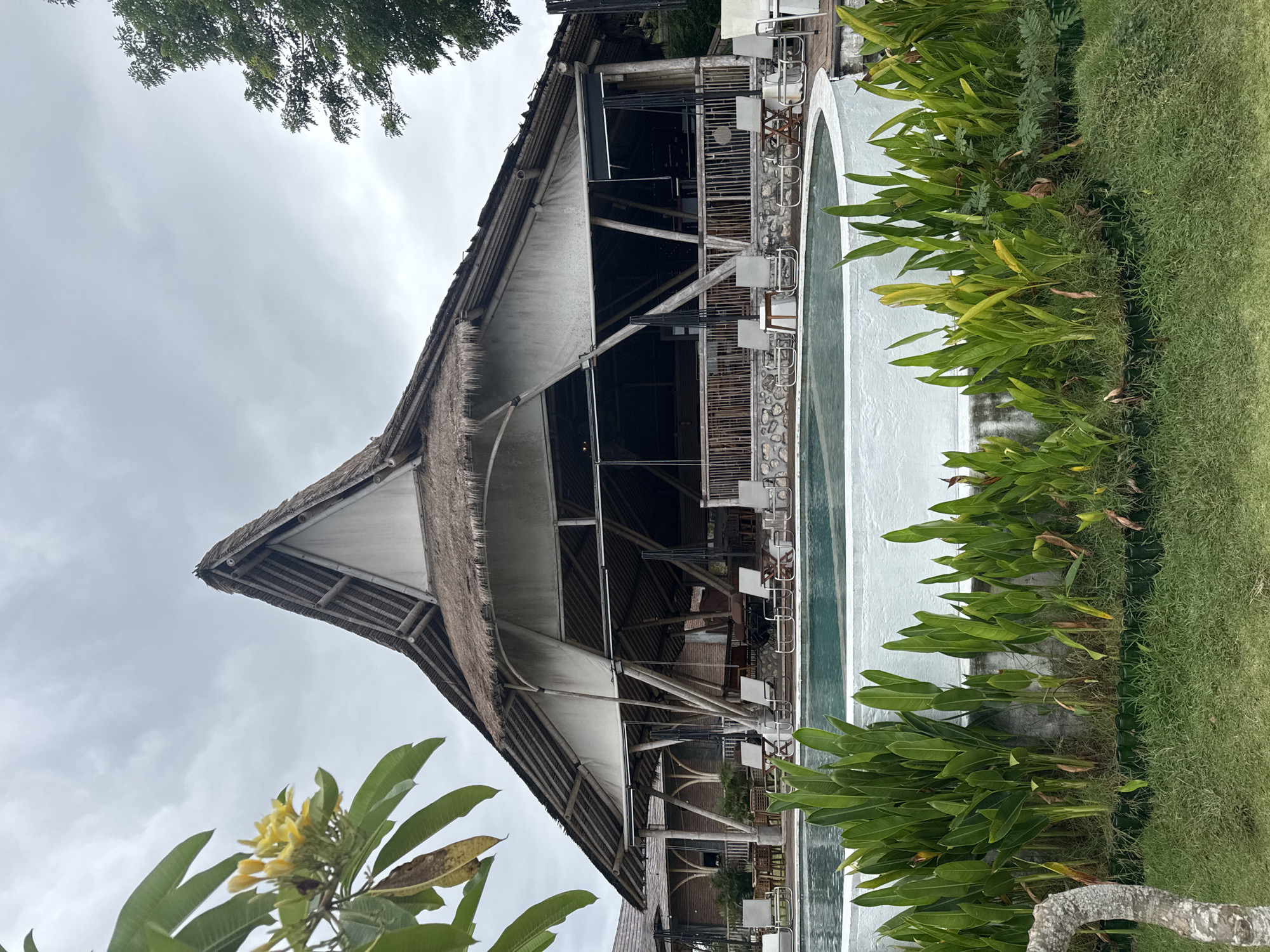 The image shows a multi-story open-air structure in Nusa Penida, Indonesia. The structure features a distinctive thatched roof and a white facade with wooden structural elements. Adjacent to the building is an elongated, presumably artificial, pool with a white border and a row of lush, green tropical plants lining its edge. A patch of grass runs alongside the plants. The sky is overcast. A flowering plant is visible in the foreground on the left side of the frame. The building is likely a resort or restaurant. The scene is devoid of people.