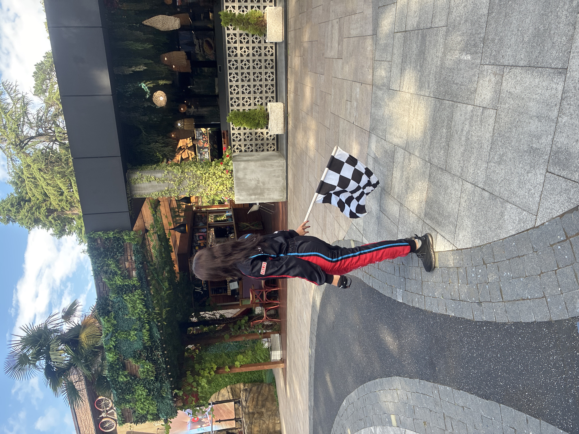 A young woman, seen from behind, stands on a paved outdoor area, holding a checkered flag. She is wearing a black racing suit with red and blue stripes down the legs and arms, and black sneakers. The flag is black and white, a symbol often used to signal the end of a race.
The setting appears to be an outdoor patio or plaza with a modern building in the background. The building has large glass windows and is adorned with hanging woven light fixtures and lush green plants, suggesting a restaurant or commercial establishment. The ground is paved with large, light-colored square tiles.
The image is taken during the daytime, with clear blue skies and sunlight casting shadows. The overall mood is celebratory, likely related to a racing event or competition. The location is identified as Sochi, Russia. No visible text is present in the image.