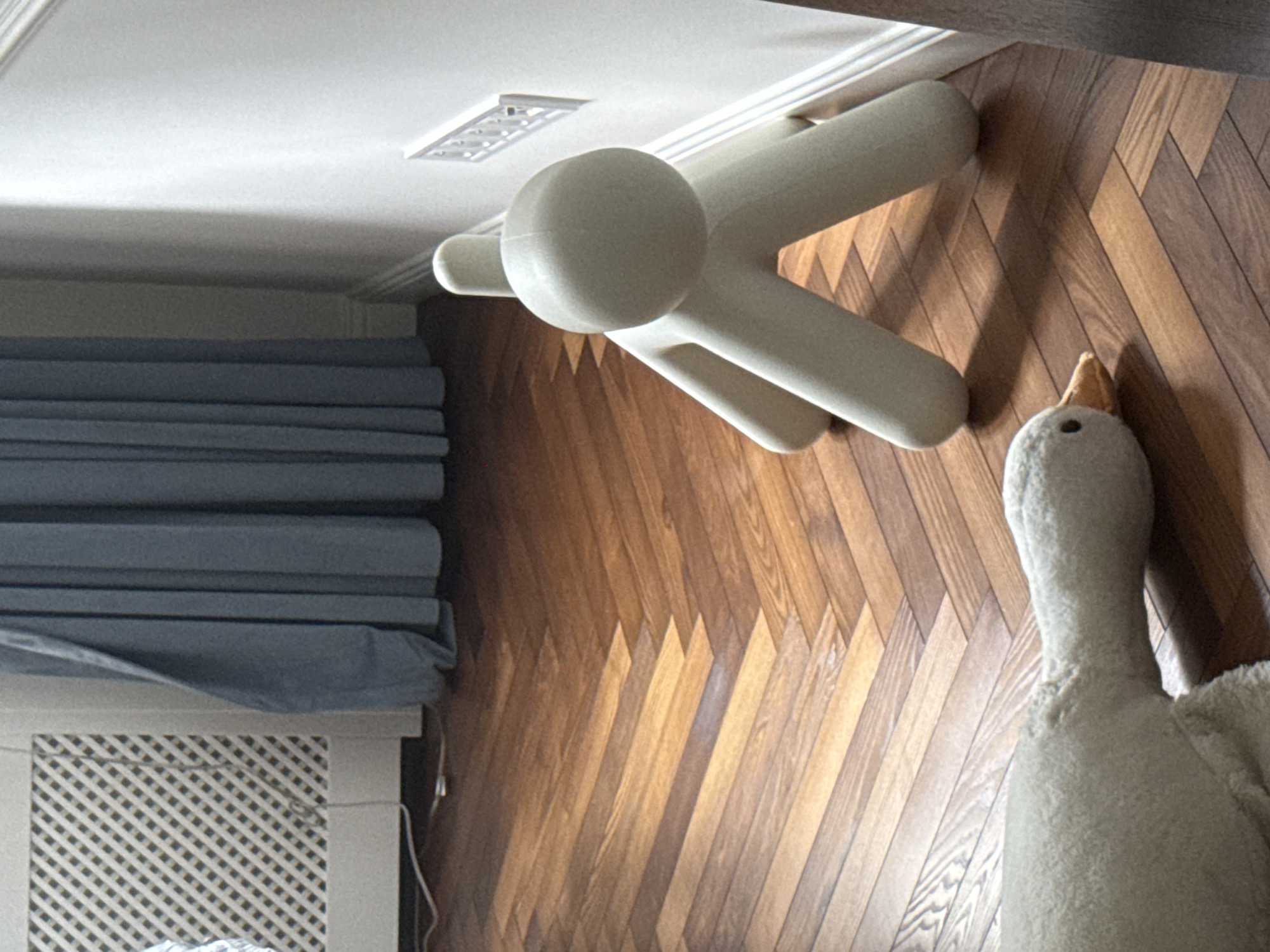 The image displays an interior scene, oriented vertically. In the foreground, a plush stuffed animal resembling a goose or swan is partially visible, with its head and neck extending upwards. The animal is a light, creamy color. Behind the stuffed animal, a wooden floor is laid in a herringbone pattern, with planks of varying shades of brown.
Further back in the image, a light-colored, abstract object with rounded shapes and cylindrical limbs is positioned on the floor. It appears to be a piece of furniture or a large decorative item. To the left of this object, a radiator cover with a white, gridded pattern is visible. A gray fabric, possibly a blind or curtain, is draped over the radiator cover and extends upwards. The ceiling is white and features a rectangular, recessed light fixture with a grid pattern. The overall impression is of a room that might be a living space or a child's room, given the presence of the stuffed animal and the playful design of the abstract object. The lighting suggests it is daytime, with natural light possibly coming from a window out of frame. There is no visible text in the image.