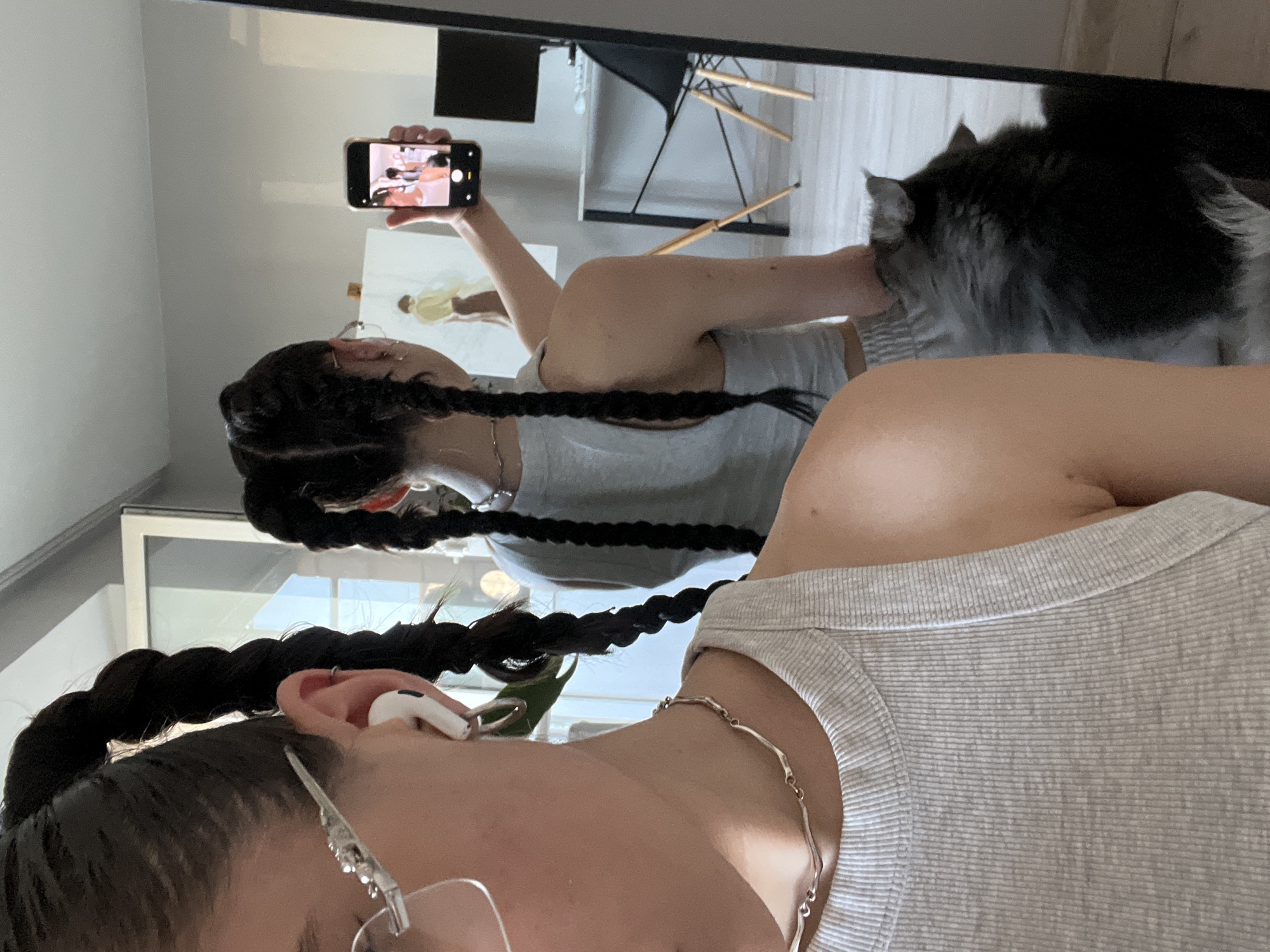 The image depicts a young woman taking a selfie in a room that appears to be her home. She is standing in front of a mirror, which reflects her and the surrounding environment. The woman has dark hair styled in two long braids, and she is wearing a grey ribbed tank top and grey shorts. She is also wearing glasses and white earbuds. In her right hand, she holds a smartphone, which she is using to take the photo. The mirror also reflects a partial view of a painting on an easel in the background, a desk with a computer monitor and a chair, and a fluffy cat sitting near the woman's feet. The room is lit by natural light coming from a window, and the overall impression is one of a casual, personal space. There is no information in the image to suggest the location is Ternopil, Ukraine.