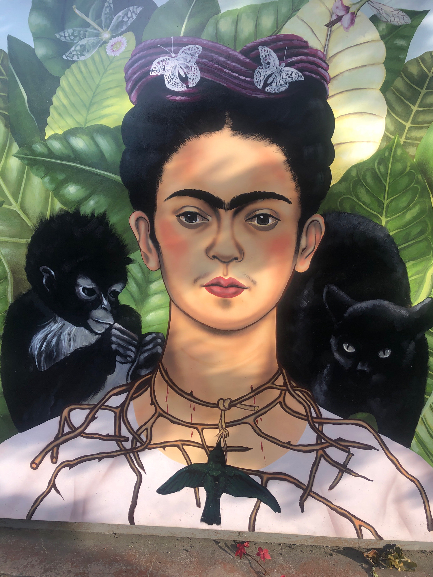 The image is a mural painted on an outdoor wall, depicting a portrait of Frida Kahlo. She is shown with her signature unibrow and hair styled in a braided bun adorned with white butterflies. Her gaze is direct and stoic. To her left, a small black monkey with white fur around its face is perched, holding its hand to its mouth. To her right, a black cat with piercing eyes is nestled behind her shoulder. Thorny vines wrap around Kahlo's neck, with blood drops on the thorns and a small black bird hanging from one of the vines. The background consists of lush, large green leaves and tropical foliage, with faint imagery of dragonflies and flowers. The mural appears to be located in an outdoor setting, possibly a public space, given the visible concrete ledge or wall base at the bottom of the image, and the slight shadows suggesting sunlight. The mural is situated in Интурист Снт, Russia.