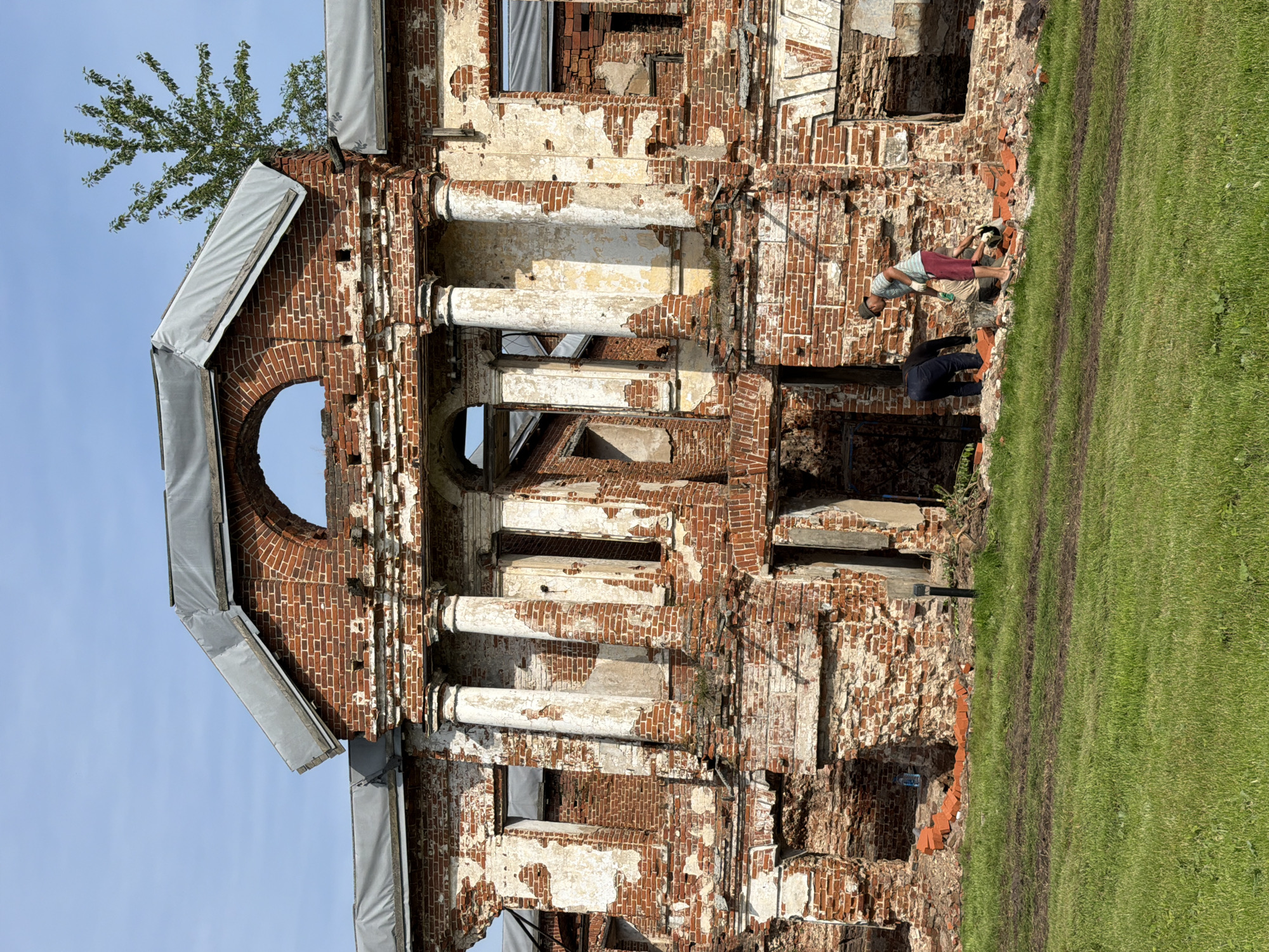 In Usolye, Russia, a partially ruined brick building stands under a clear blue sky. The structure exhibits significant decay, with exposed brickwork and missing plaster, revealing its aged construction. A large arched window is visible at the top of the central facade, framed by columns. In front of the building, on a grassy area, three individuals are engaged in what appears to be restoration or demolition work. One person, wearing a striped shirt and red shorts, is kneeling and holding a trowel, working with loose bricks. Another person, dressed in dark pants and a dark shirt, is also bending over near the bricks. A third individual, wearing a light-colored shirt and dark shorts, stands beside them. Scattered bricks are present on the ground around the workers and in front of the building. The environment is sunny, with green grass covering the foreground and the immediate area surrounding the building.
