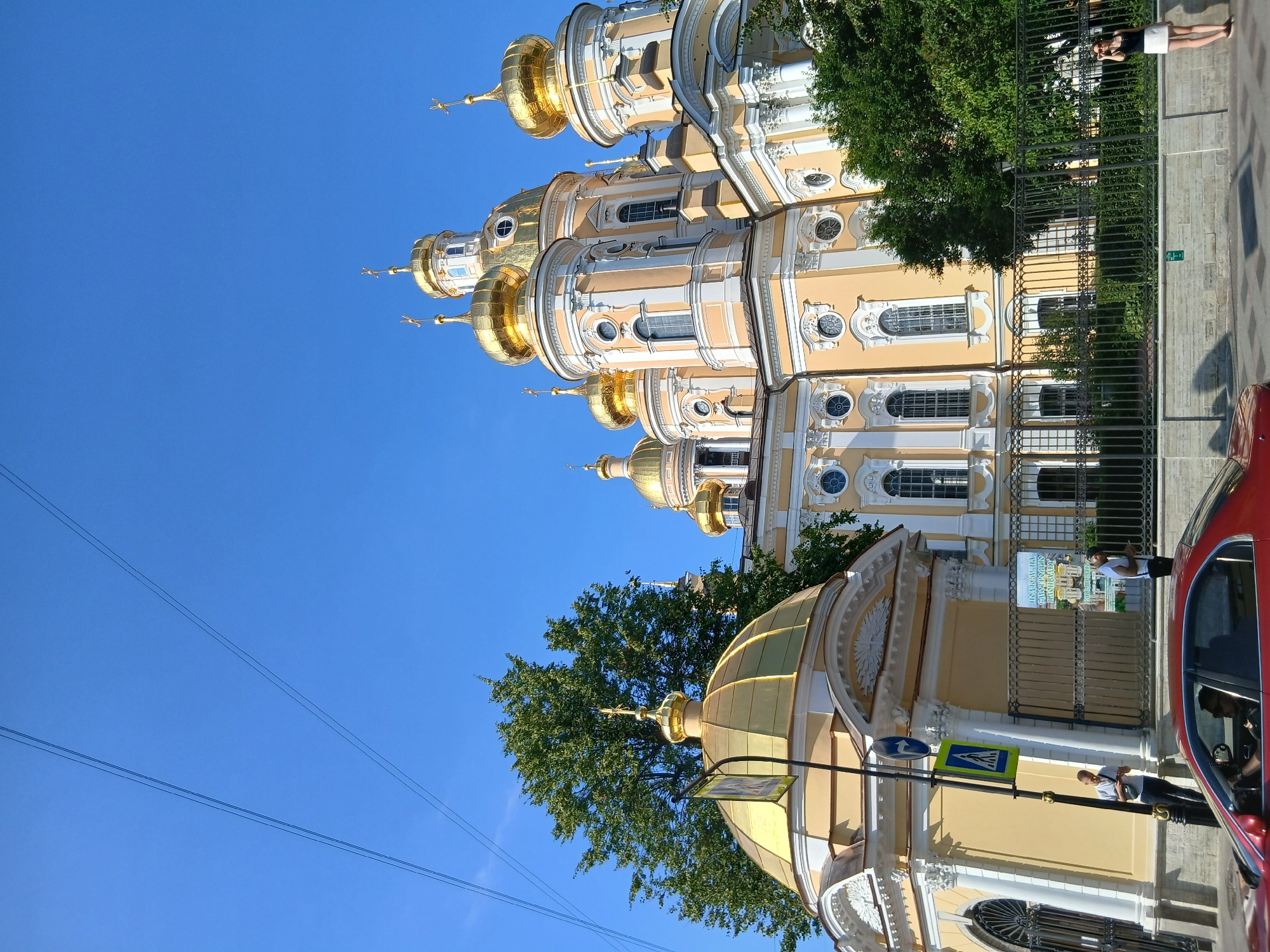 This is a vertical, eye-level, outdoor photograph of a large, ornate, yellow building with multiple golden domes. The building appears to be a church or cathedral. The architecture is classical with columns, arches, and decorative moldings. Several of the domes have crosses on top.
In the foreground, to the right, a portion of a red car is visible. Behind the car and a black metal fence, there are people walking on a sidewalk. One person, a woman wearing a white skirt and dark top, is standing by the fence looking to the left. Another person, a man, is standing next to a traffic sign.
A large green tree with dense foliage is visible behind the fence, partially obscuring the building. The sky is a clear, bright blue, suggesting a sunny day. There are overhead wires in the lower left portion of the image.
The image is taken from a street-level perspective. The overall impression is one of a bright, sunny day in a city with significant historical architecture. There is no visible text in the image.