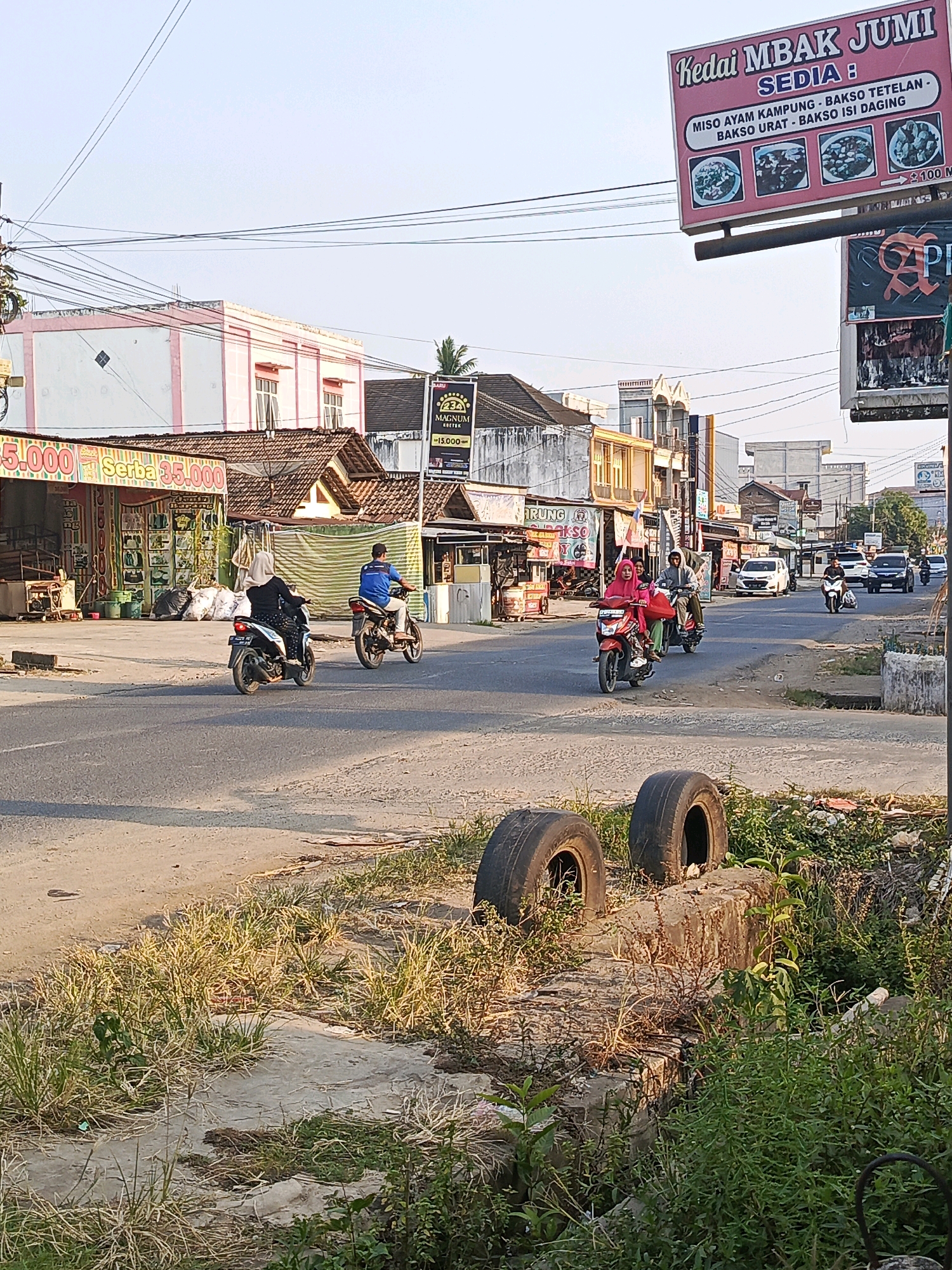 The image depicts a street scene in Megang Sakti II, Indonesia, under a clear sky. A red sign for "Kedai Mbak Jumi" is prominently displayed, advertising "Miso Ayam Kampung, Bakso Tetelan, Bakso Urat, Bakso Isi Daging". Below this, another sign reads "23A Magnum Kopi".
The street is paved with asphalt and shows signs of wear. Several motorcycles are in motion. On the left, a person on a black motorcycle wearing a grey jacket and black headscarf is traveling away from the camera. To their right, a person on a blue motorcycle, also traveling away, wears a blue shirt and a dark helmet. In the center, a person on a red motorcycle, wearing a pink headscarf and a pink jacket, is moving towards the camera, accompanied by a passenger in a grey hoodie. Further down the road, other vehicles, including a white car and several motorcycles, are visible.
Along the sides of the road, there are buildings, some with tiled roofs. On the left, a long, low building with a pink and white facade has banners displaying prices like "35.000". On the right, the foreground features overgrown grass, weeds, and two discarded tires on top of a concrete structure. The overall impression is of a busy, developing urban environment with elements of everyday life unfolding on the street.