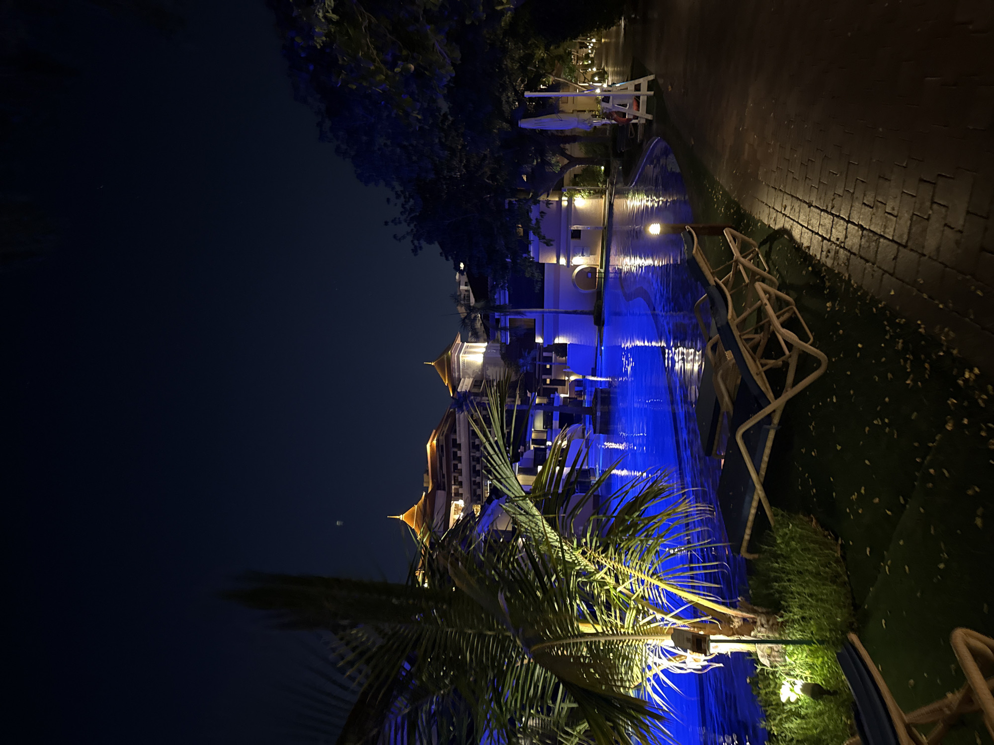 The image depicts a nighttime scene at a luxury resort in Dubai, United Arab Emirates. In the foreground, a palm tree with illuminated fronds stands to the left, casting a green glow. Adjacent to the palm tree are several empty lounge chairs, positioned on a grassy area. A paved walkway, made of dark bricks, runs along the right side of the image, illuminated by lampposts.
The focal point of the image is a large swimming pool, its water illuminated by vibrant blue underwater lights, creating a striking contrast with the dark night sky. The pool reflects the lights and the surrounding architecture.
In the background, a multi-story resort building with distinct golden-capped towers is visible. The building is illuminated by warm yellow lights, highlighting its intricate architectural details. Other structures and trees are also present, contributing to the resort's ambiance. The sky is a deep, dark blue, with a faint crescent moon visible near the top. The overall atmosphere is serene and luxurious.