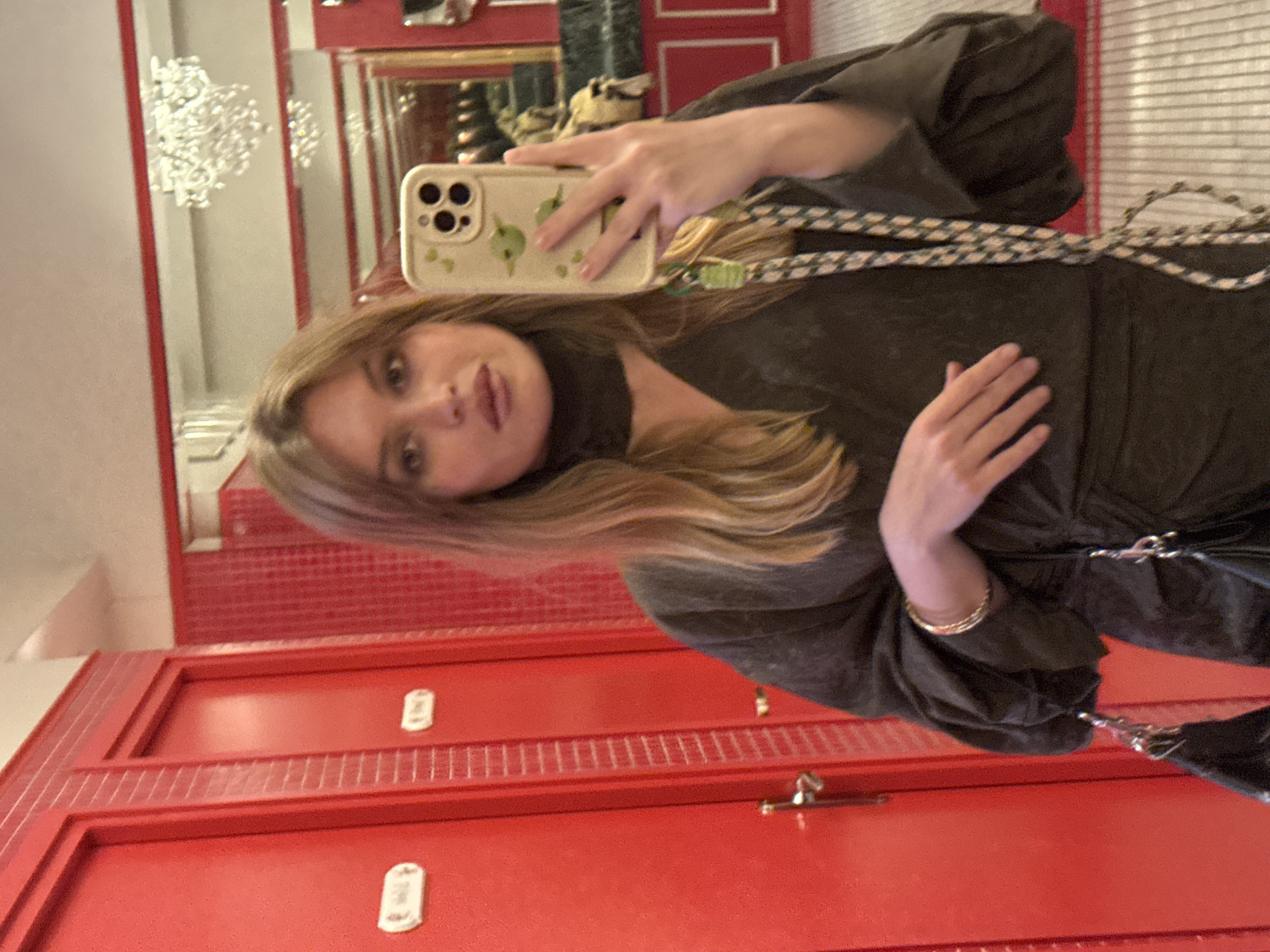 A young woman is taking a selfie in a restroom in Paris, France. She is holding a phone with a cream-colored case with leaf designs. She wears a dark-colored dress with sleeves and a dark strap. She has light brown hair. The background consists of red doors with silver numbers and hardware. A chandelier is reflected in the mirror behind her.
