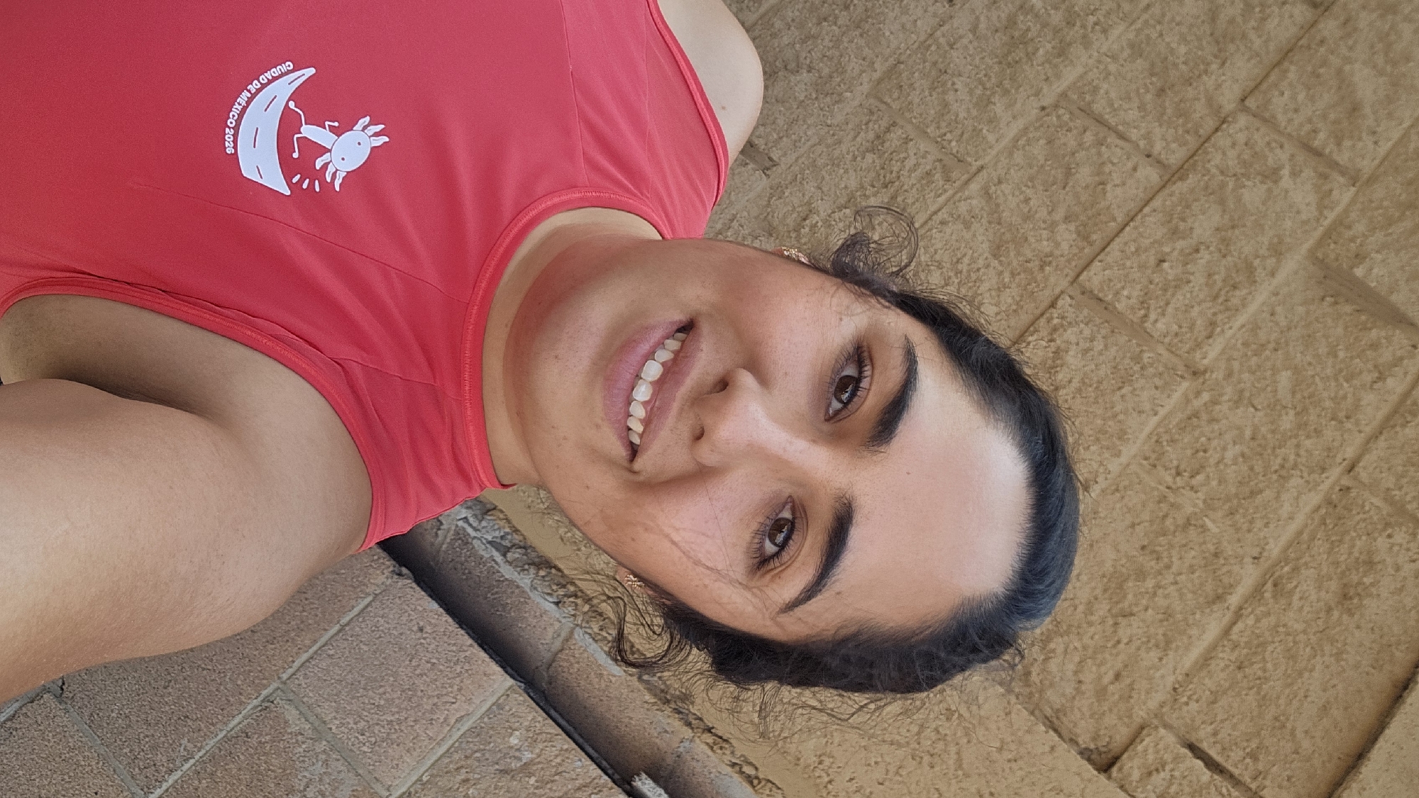 A young woman with brown eyes and dark hair is posing for a selfie outdoors. She is smiling and wearing a red tank top with a white logo on the upper left side that reads "Ciudad de México 2026" along with a cartoon-style image. The background is a light brown brick wall. The location is likely San Bernardino Tlaxcalancingo, Mexico, based on the image context. The time of day appears to be daylight.