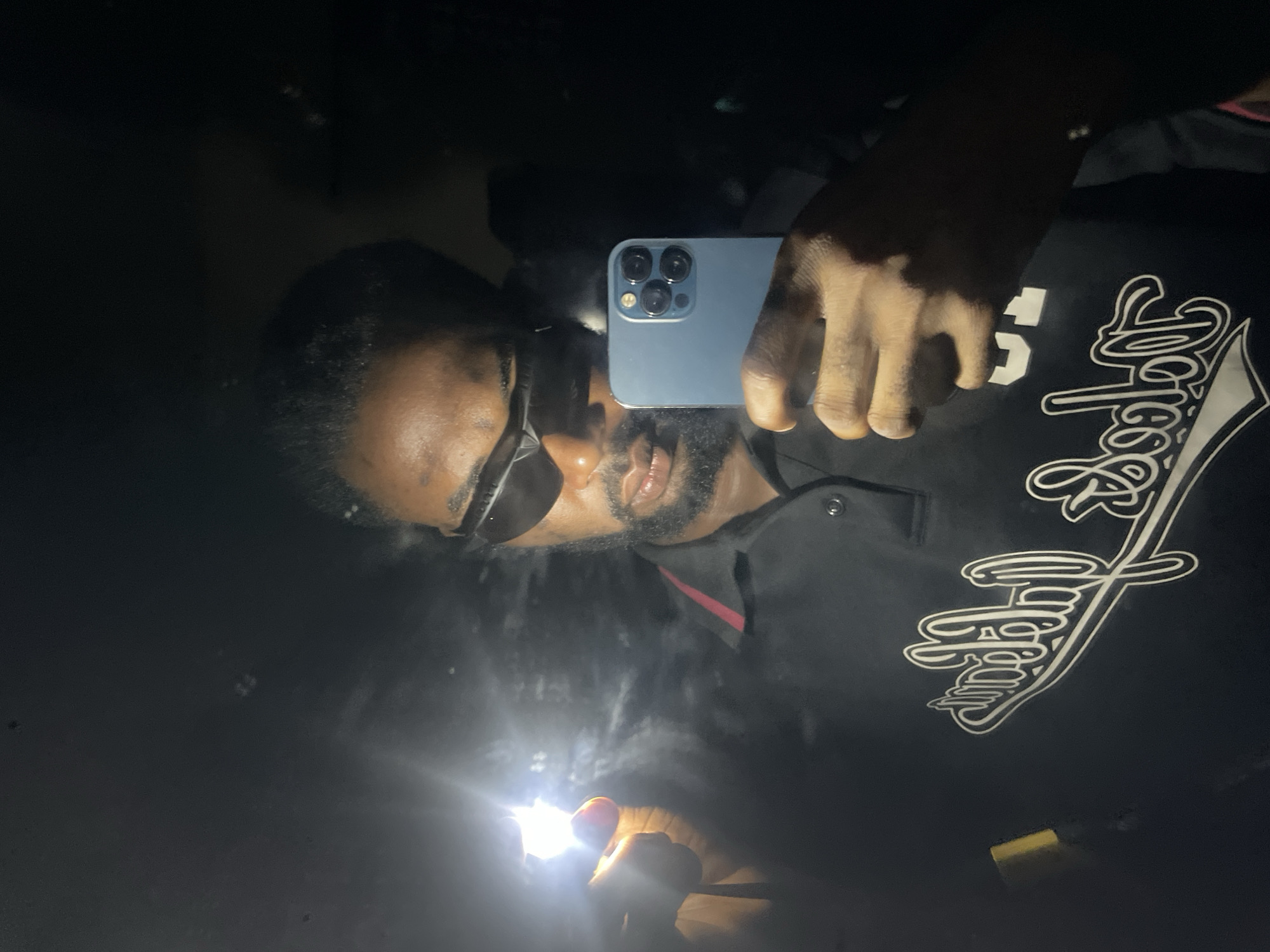 An adult male is depicted in a mirror selfie taken in a low-light environment. The subject has dark skin and a beard, and is wearing dark, wrap-around sunglasses and a black polo shirt with a red accent on the collar. The shirt features a prominent white graphic in a stylized script, which includes text resembling "Lokoja." The subject holds a light blue smartphone, identified by its triple camera array (consistent with an iPhone 12 Pro or 13 Pro model), in his right hand, directed towards the reflection. A bright white light source is held in his left hand, producing a strong light flare on the left side of the image. The background is dark and indistinct, consistent with a reflective surface in a poorly lit space. While the shirt prominently displays text referencing "Lokoja," a city in Nigeria, the precise location within the city is not discernible from the visual information provided in the image.