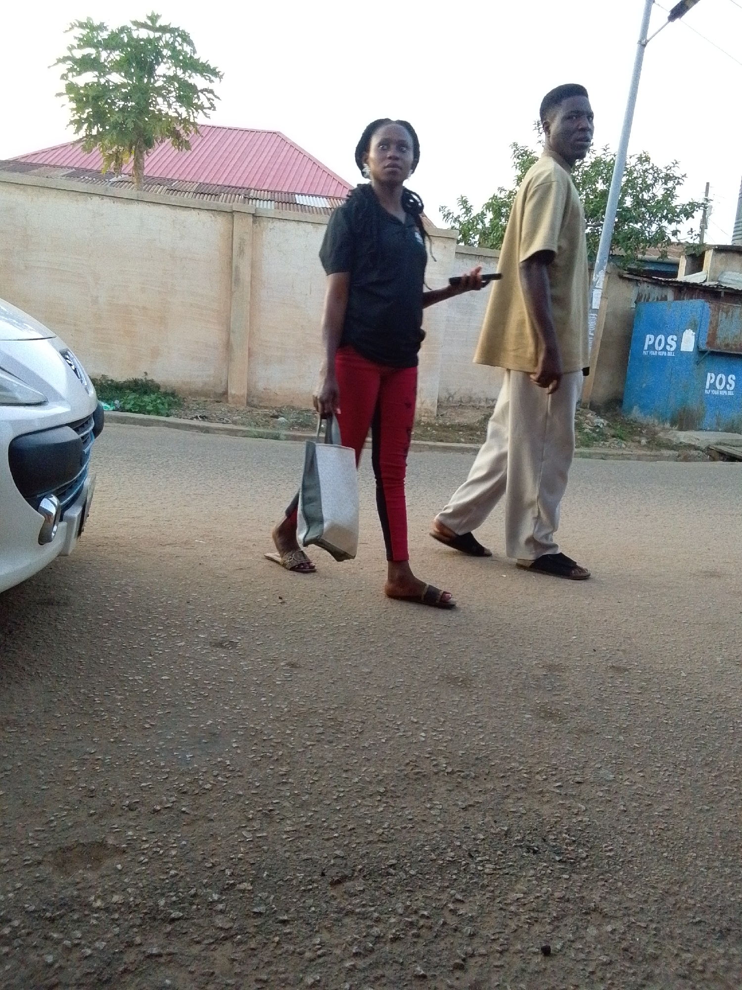 Two individuals, a woman and a man, are captured walking on a textured asphalt road, likely in a street scene during late afternoon or early evening. The lighting is bright but not harsh, suggesting the sun is lower in the sky.
The woman is positioned on the left, slightly ahead of the man. She has dark skin, wears a black short-sleeved top, red leggings, and brown flat sandals. Her hair is styled in dark braids. She carries a light-colored (white/grey) shopping bag in her left hand and appears to be holding a black smartphone in her right. She looks intently towards the viewer or slightly past the camera.
The man is behind her on the right, also with dark skin and short dark hair. He wears a loose-fitting, light brown (khaki) short-sleeved shirt, light-colored (beige) trousers, and dark flat sandals. He is looking back towards the camera as he walks.
To the far left, the front portion of a white car, possibly a Peugeot given its grille design, is visible. The background features a tall, light-colored wall with visible texture, topped by corrugated metal sheeting and a red corrugated roof. A small tree with green leaves is seen above the wall and roof on the left. On the right side of the background, there's a light pole and two blue kiosks or boxes.
The blue boxes clearly display the text "POS" and "PAY YOUR NEPA BILL," indicating a Point of Sale service often associated with bill payments in Nigeria. The road surface is composed of small stones and gravel embedded in asphalt.