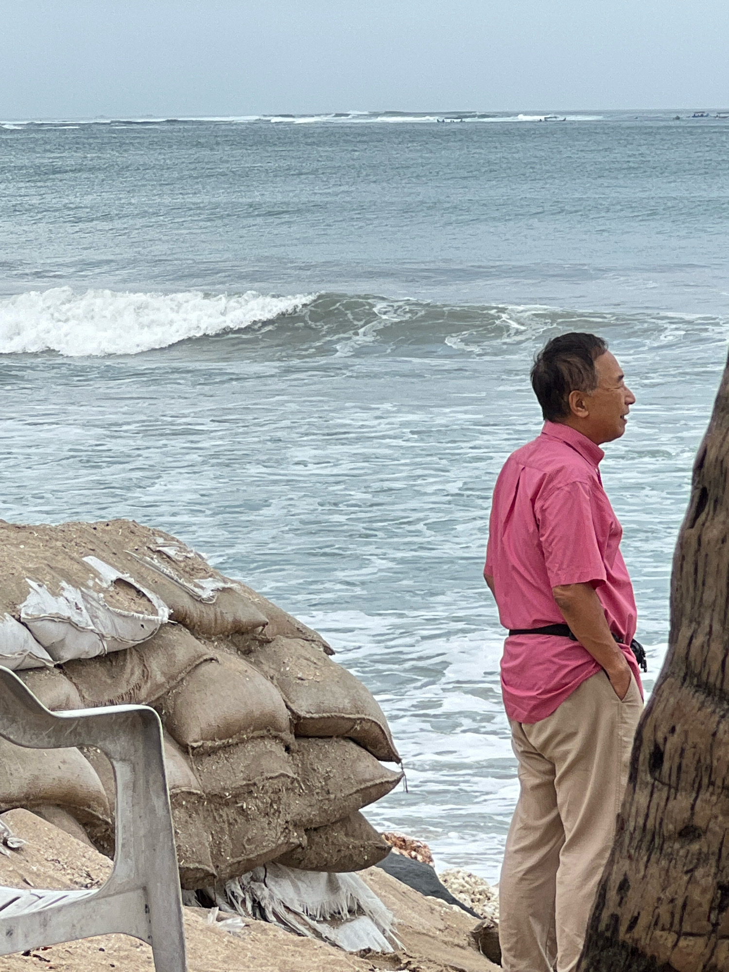 An adult male, identified as East Asian, stands on a beach in Kuta, Indonesia. He is wearing a pink short-sleeved collared shirt tucked into tan trousers. His hands are in his pockets, and he is looking out at the ocean. The ocean is choppy with waves breaking. Behind him, there are sandbags stacked as a barrier against the water. To his left, a metal chair is partially visible. To his right, the trunk of a large tree obscures part of the scene. The sky is overcast. Several small boats are visible on the horizon in the distance.