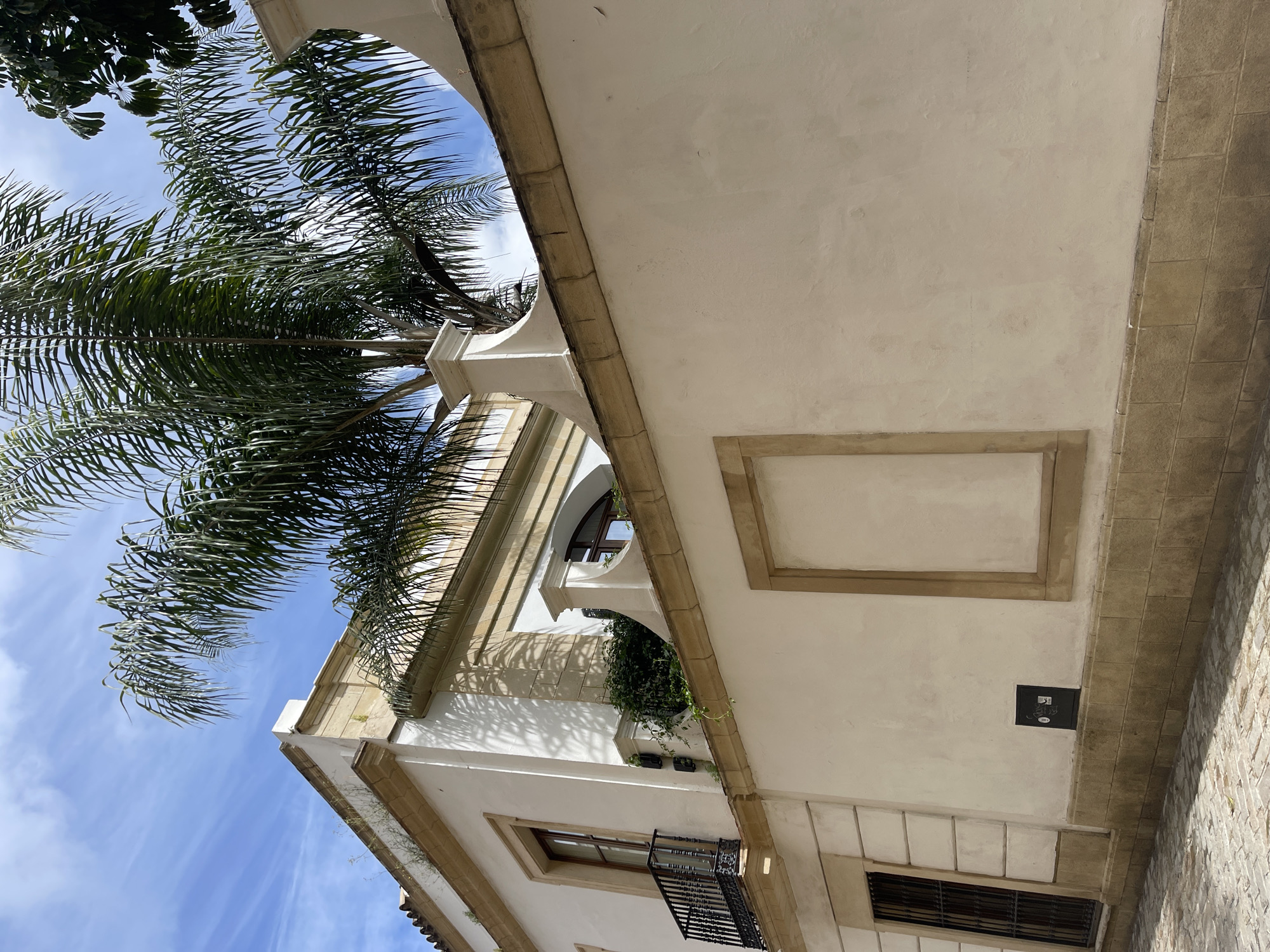 Captured from a low angle, the photograph showcases the architectural details of a building in Jerez De La Frontera, Spain, juxtaposed with a vibrant palm tree against a blue sky. The building's facade is characterized by its cream-colored walls, framed windows, and an archway, which suggests a courtyard or passageway. The palm tree's fronds extend into the frame, adding a tropical touch. The sky is partially visible between the palm leaves, revealing light cloud formations. A small black box or fixture is noticeable on the wall near the building's edge.