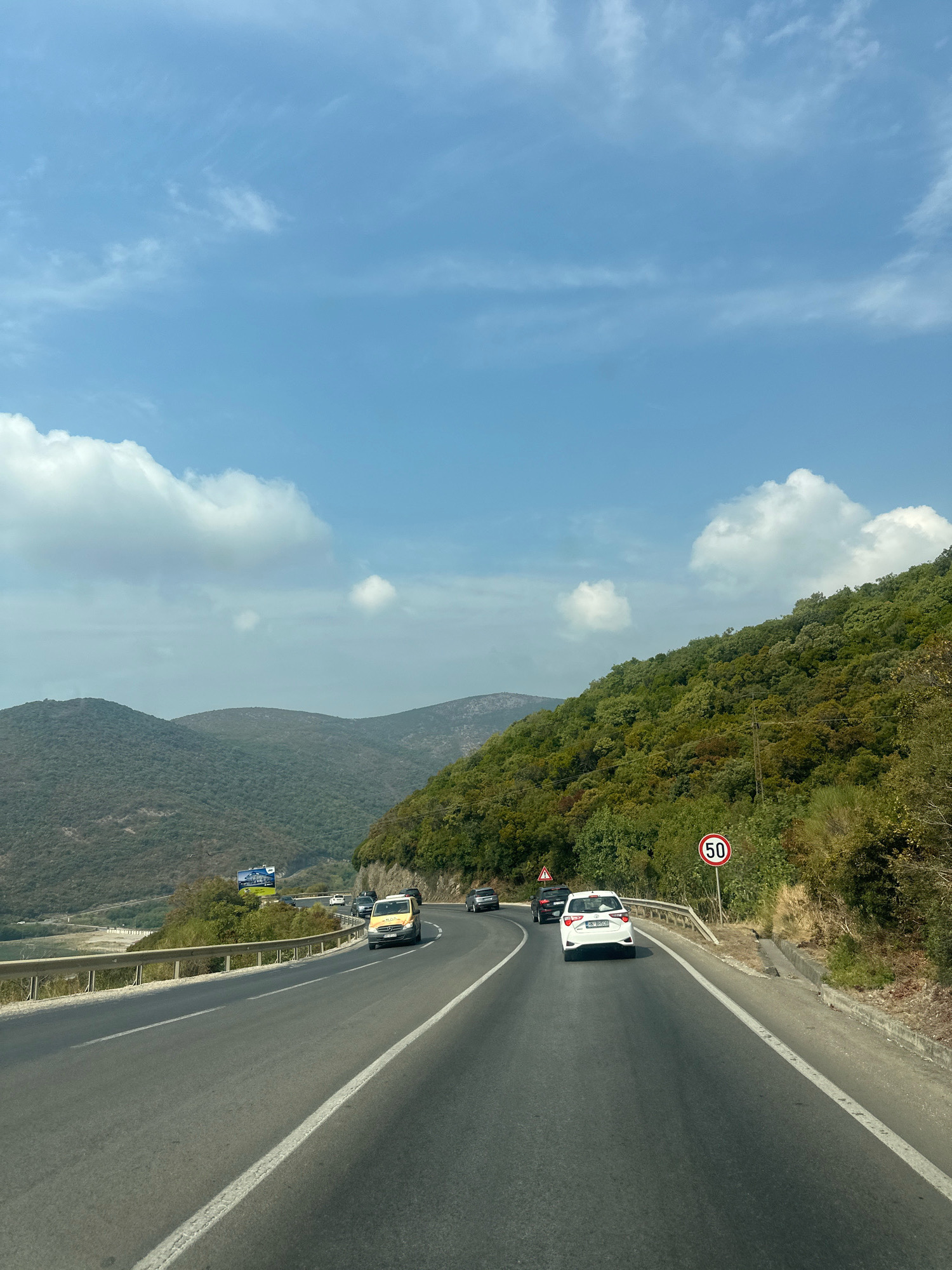 A scenic road winds through lush green hills under a partly cloudy blue sky. Several cars are driving on the road, which has a guardrail on the left side and a concrete barrier on the right. A speed limit sign indicating "50" is visible on the right side of the road. The landscape suggests a mountainous or hilly region, potentially near a body of water which is visible in the distance on the left. The overall impression is a travel scene on a sunny day.