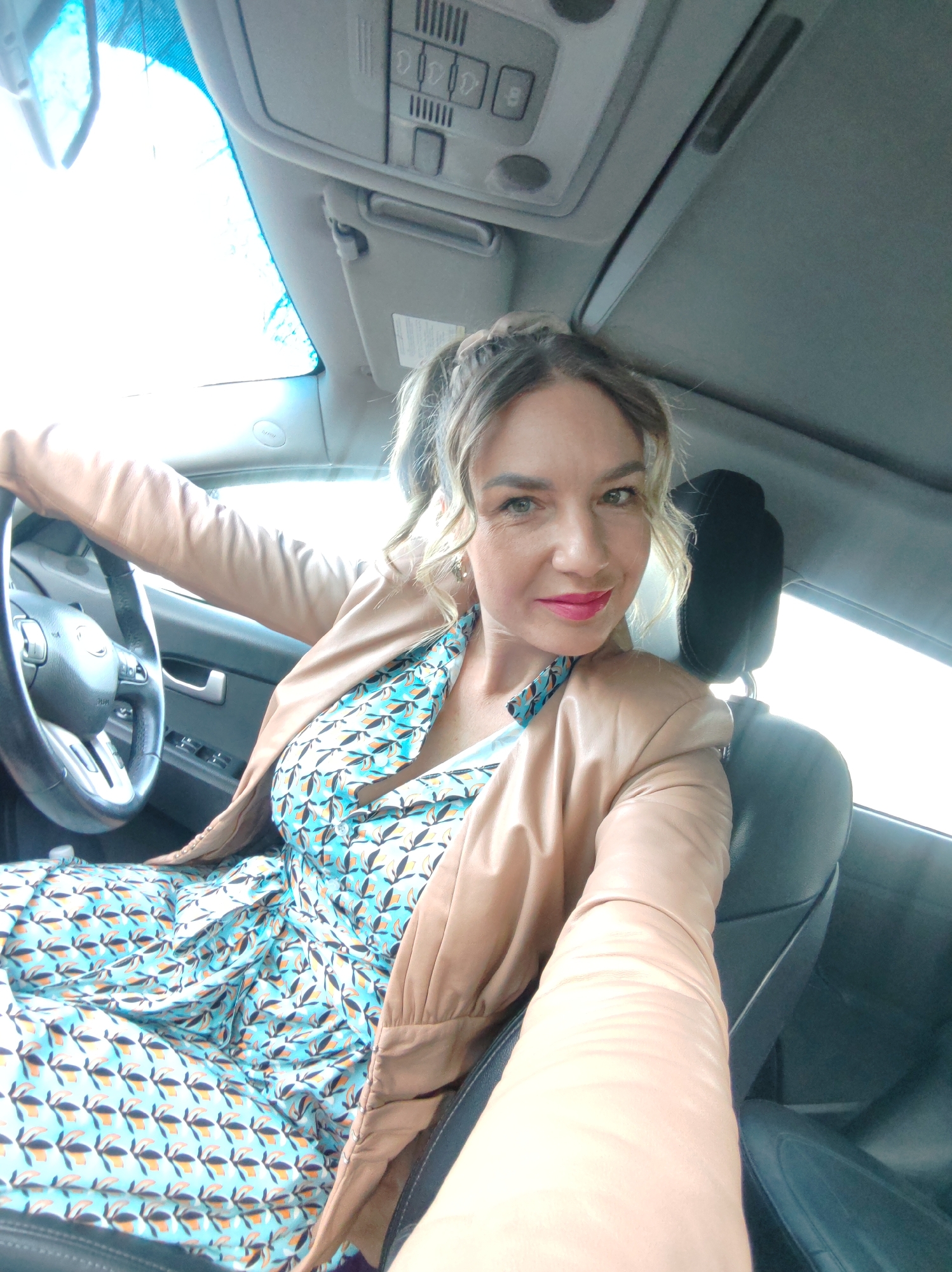 A woman is sitting in the driver's seat of a car. She is wearing a light brown leather jacket over a blue and white patterned dress. She has blonde hair pulled back in a ponytail and is wearing bright pink lipstick. She is holding the steering wheel with her left hand and extending her right arm towards the camera. The car is parked, and the interior is visible, including the dashboard, the headrest of the passenger seat, and the car's ceiling. The scene appears to be inside the car, during the daytime, with natural light filtering through the windows. Location is unknown, not identifiable as Petropavlovsk-Kamchatsky, Russia.