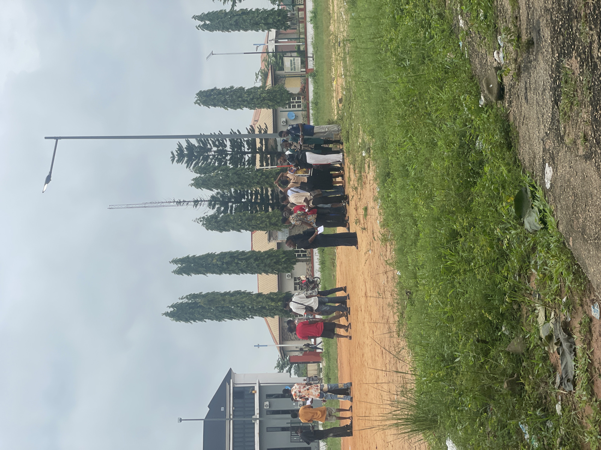 In Ihiagwa, Nigeria, a group of approximately 12 individuals, both male and female, are gathered outdoors on a dirt surface. The group appears to be engaged in a discussion or instruction, with some holding papers. They are standing near a light pole and several tall, slender trees. In the background, there are buildings, including a modern-looking structure with dark roofing and a longer, single-story building with a reddish-brown roof. A sign that reads "PUBLIC PROSECUTION DIVISION REGISTRY CENTER OFFICE" is visible on one of the buildings. The ground is a mix of dirt and patches of green grass, with some visible debris. The sky is overcast.