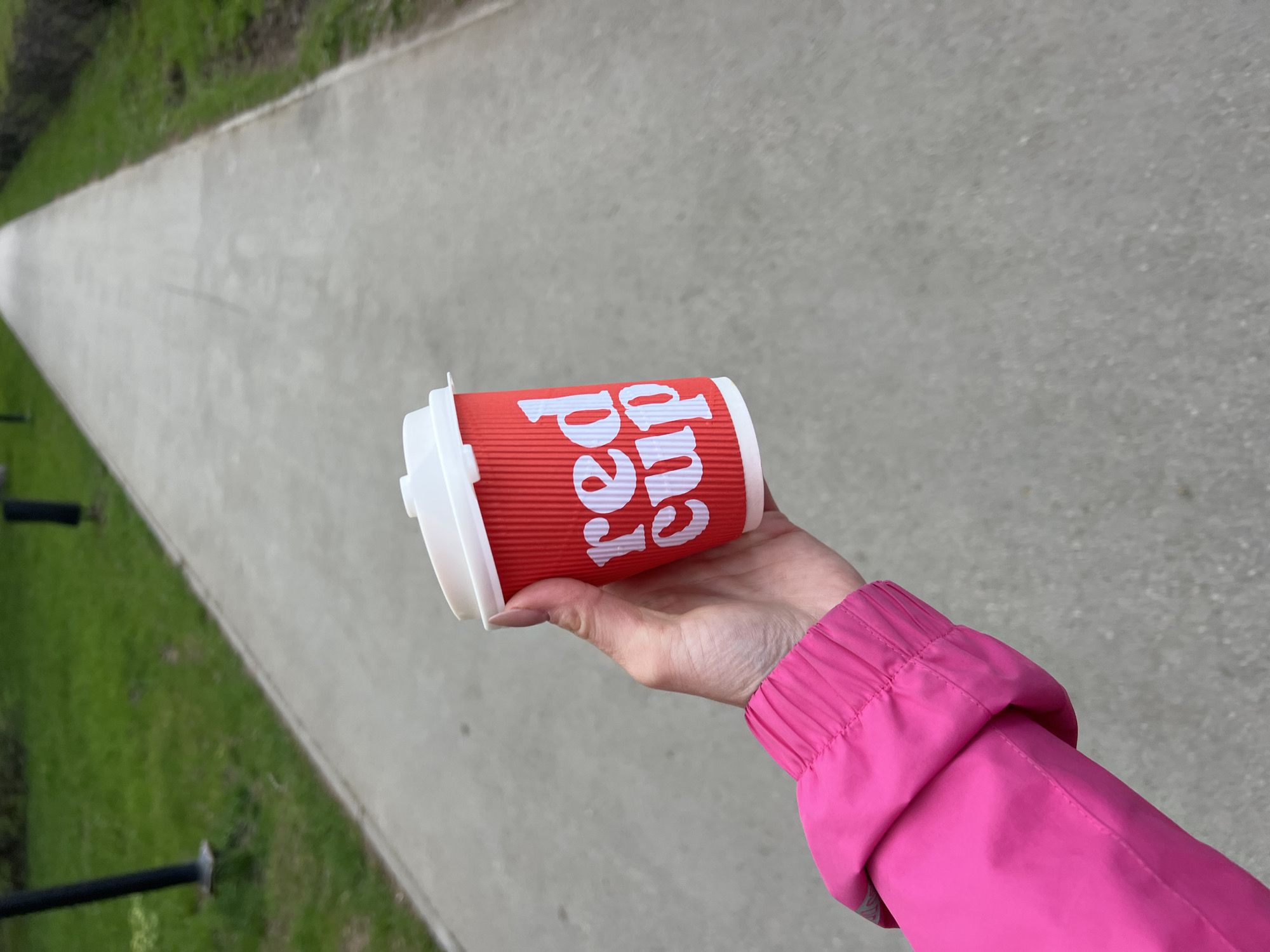 A red paper coffee cup with a white lid is held in a person's hand against a grey concrete path, the cup is labeled "Red Cup." The person is wearing a bright pink jacket. The path extends into the background with greenery visible on the left edge. The photo appears to be taken outdoors, potentially in a park in Perm, Russia.