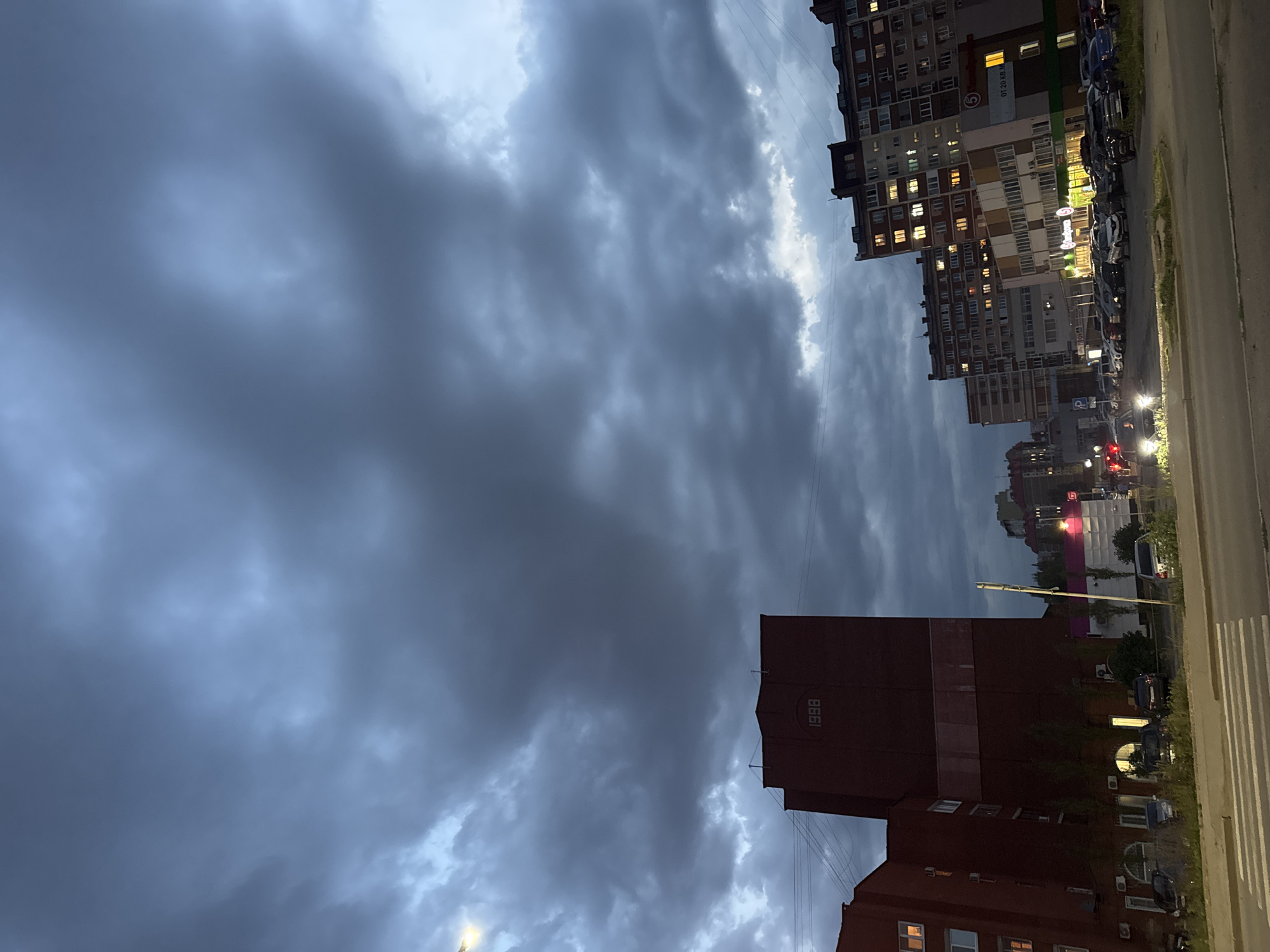 The image depicts a cloudy sky over a city scene at dusk. In the foreground, a crosswalk is visible on a paved road. To the left, a dark red brick building with several lit windows rises up, bearing the number "1998" in white lettering on its upper facade. To the right, several multi-story apartment buildings are lit from within, with a commercial building featuring illuminated signage at street level. A parking lot filled with various cars is situated between the buildings and the road. Trees and other greenery line the area in front of the buildings. The overall impression is of an urban environment at the end of the day, under a dramatic, overcast sky. The presence of Russian text on one of the signs suggests the location is within Russia. While the specific location within Ufa cannot be definitively determined from the image alone, it appears to be a residential and commercial area.