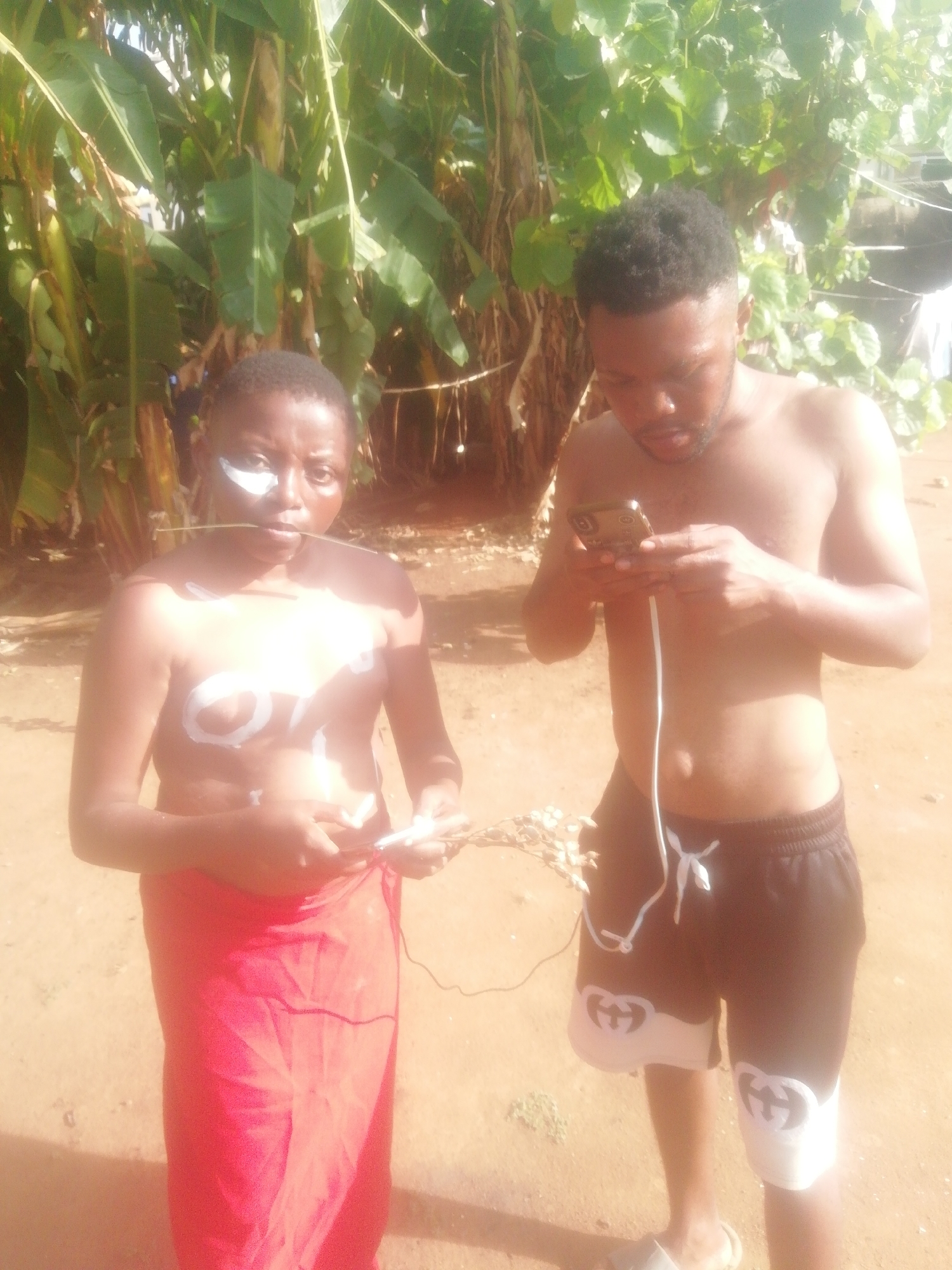 An outdoor, sunlit image features two individuals, a female and a male, standing on sandy ground in front of lush green foliage, likely tropical plants such as banana trees.
The female, positioned on the left, has dark skin and short dark hair. She looks directly at the camera with a neutral expression. White markings are painted on her face around her right eye, and stylized white letters or shapes, possibly reading "OYI" or "CVI," are painted across her bare chest. A thin, light-colored stick or twig is held in her mouth. She wears a red fabric wrap around her lower body and holds a small, white object in her hands. A white cable is visible near her hands, extending towards the male.
The male, on the right, is shirtless and has short dark hair and a beard. He is absorbed in a smartphone he holds with both hands, looking down at the screen. A white cable hangs from his phone, trailing downwards. He wears black shorts with white trim and prominent white "GG" logos, resembling the Gucci brand, on the lower outer thigh of each leg. Light-colored flip-flops are on his feet.
The scene is brightly illuminated by natural sunlight, indicating a clear, sunny day. The ground is dry and appears to be dirt or sand. The image presents a blend of cultural aesthetics, possibly traditional body art and attire on the female, juxtaposed with modern technology and branded clothing on the male, connected by the shared white cable.