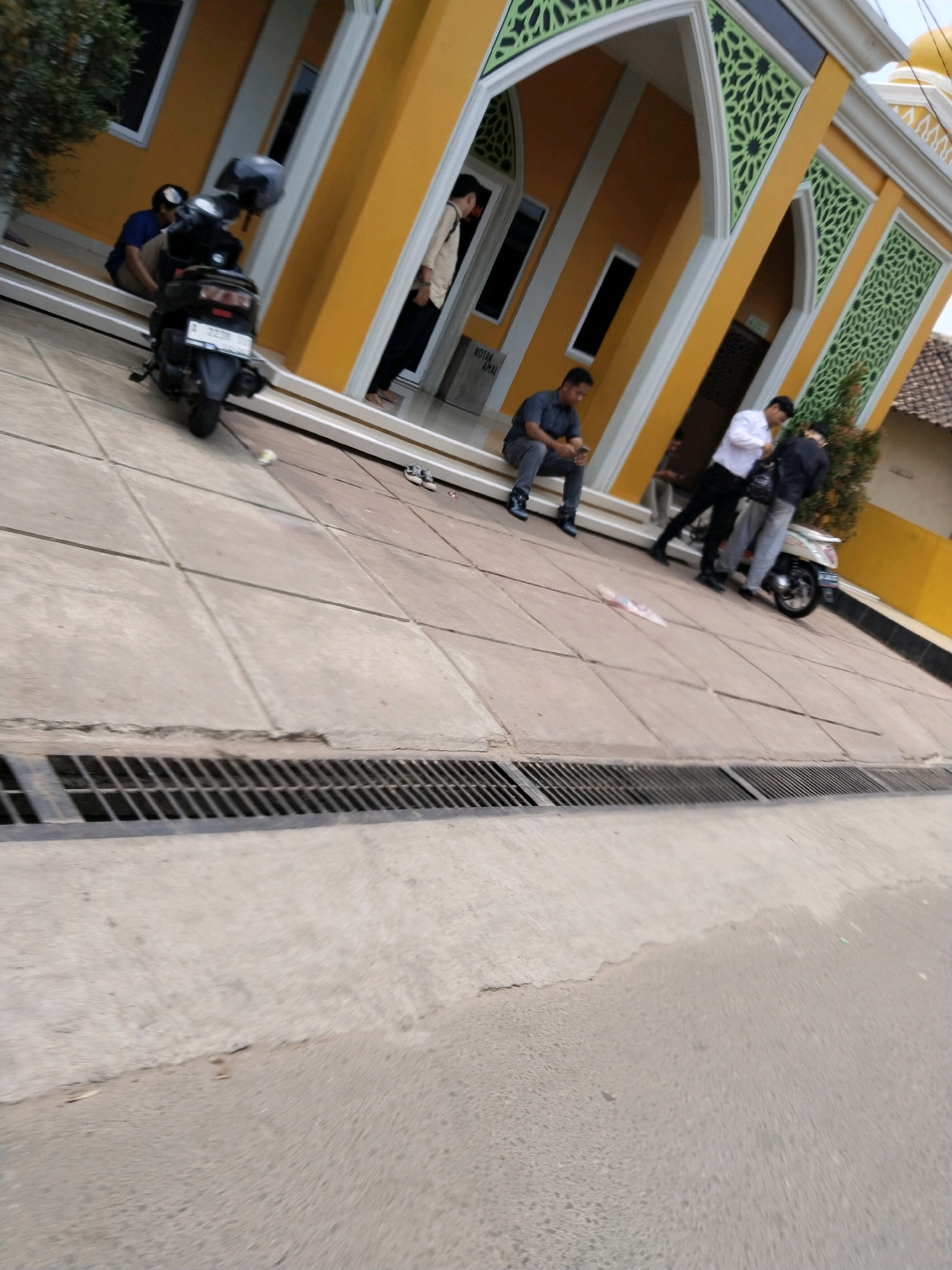 The image captures a scene in front of a yellow building with green decorative accents, likely a place of worship or public building. In the foreground, a black motorcycle is parked on a tiled walkway. To the left, a person in a blue shirt sits on a step. Further into the image, three men are standing near the entrance of the building. One man, wearing a dark shirt and light pants, is seated on the steps. Another man, dressed in a white shirt and black pants, stands next to him, looking down. A third man, in a black jacket and light-colored pants, is also present, with his back to the camera. The building's architecture suggests it may be located in a Southeast Asian context, possibly Indonesia given the provided location information regarding Serang. The overall environment appears to be an outdoor public space with a paved walkway and drainage grate.
