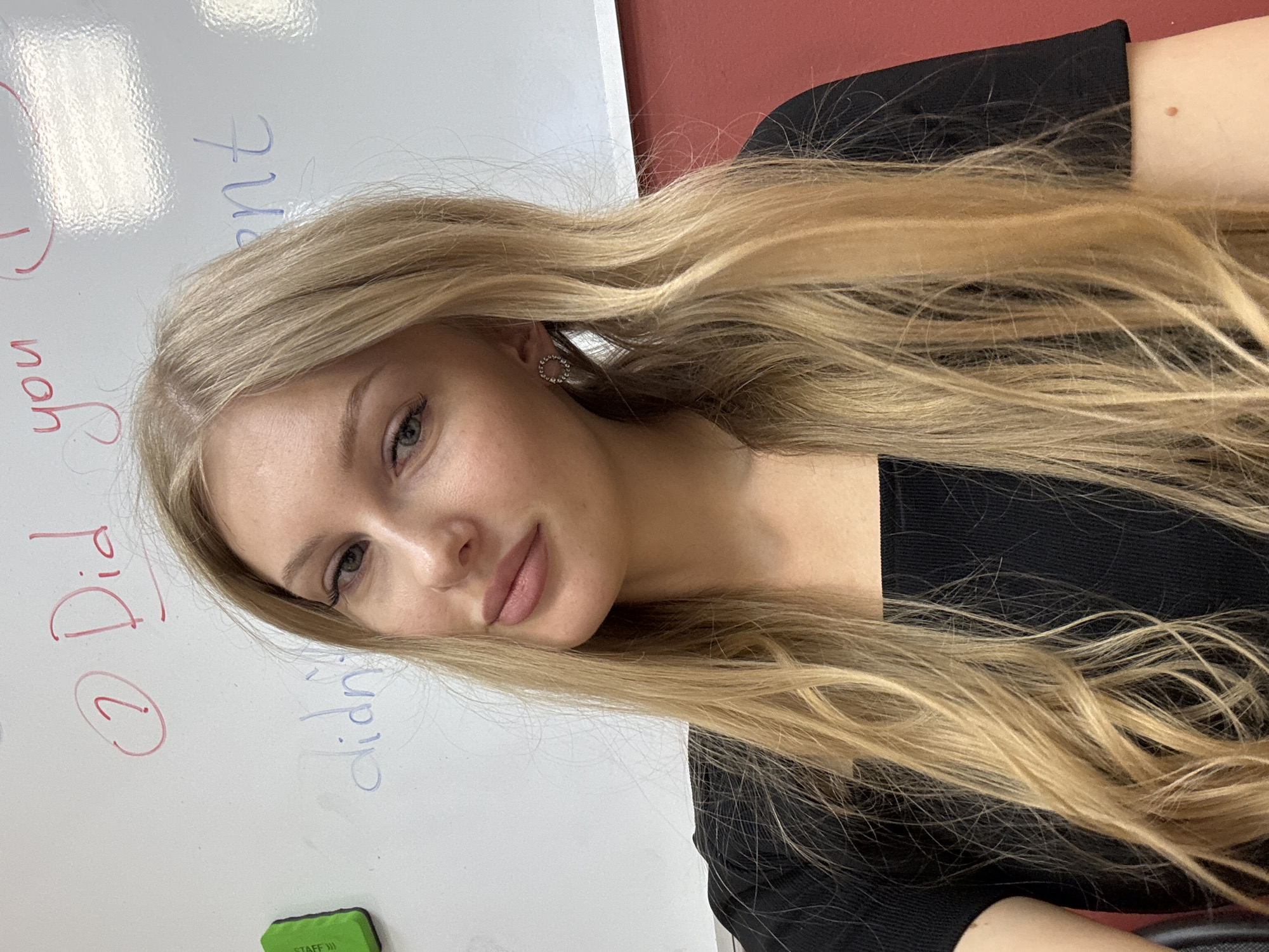 A young woman with long, blonde hair, wearing a black top and a circular earring is facing the camera. Behind her is a white board with red and blue writing, the first two words saying "Did you...". To the side of the board there is a green eraser, and partially visible, a red wall. The location is possibly a classroom in the city of Perm, Russia.