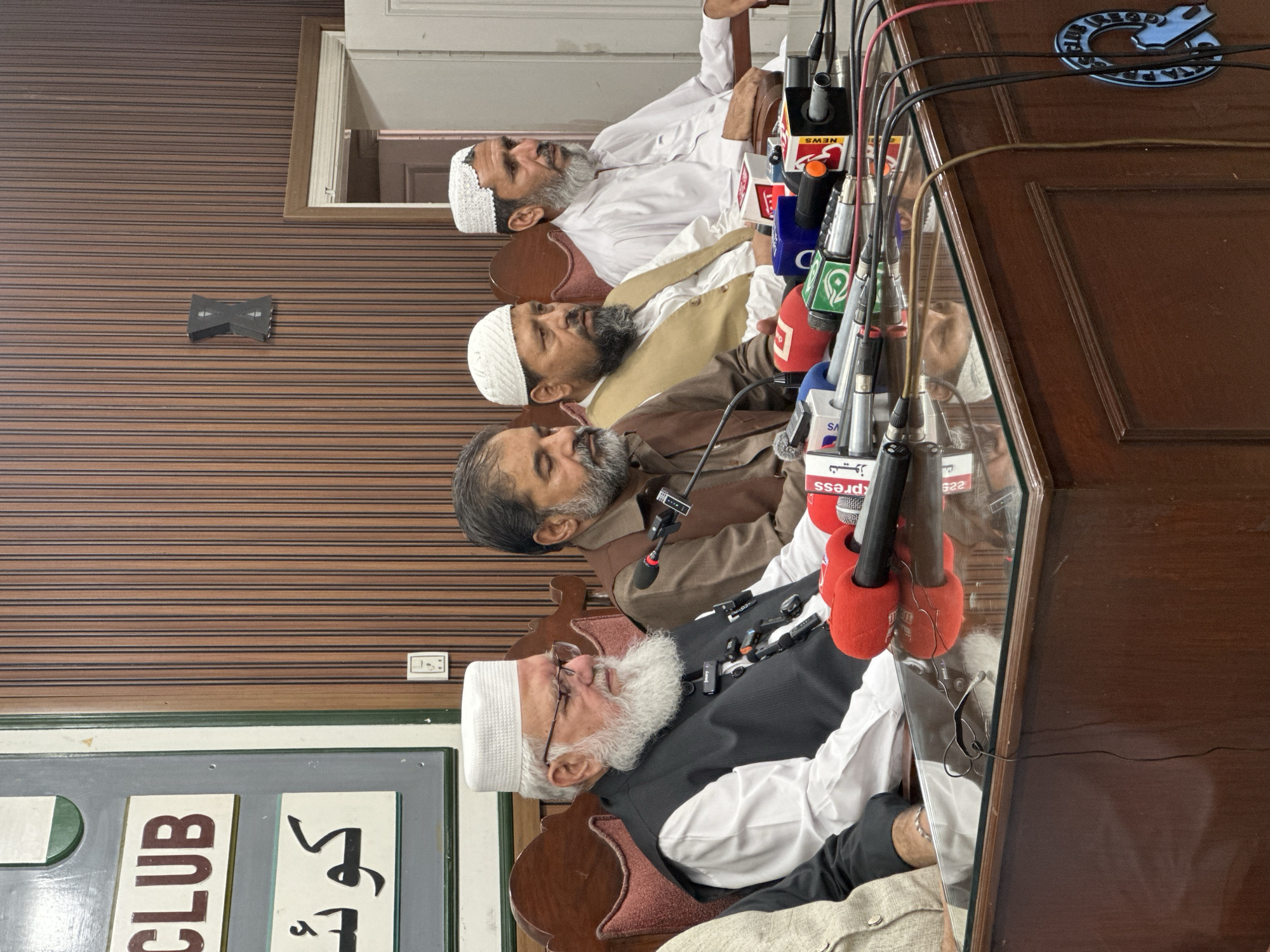 Four men are seated at a wooden desk in a room with vertically paneled wooden walls. The man on the far left is wearing a white turban and a black vest over a white shirt. He has a long white beard and glasses. The man next to him is wearing a brown shirt and a tan vest. He has a short beard. The third man from the left is wearing a white cap and a white shirt. He has a short beard. The man on the far right is also wearing a white cap and a white shirt. He has a short beard. The desk is covered with microphones and cables. A sign with Arabic script and the word "CLUB" is visible on the wall behind the men. The overall scene appears to be a press conference or a meeting. Based on the sign containing "Q CLUB" and the context of "Quetta City Tehsil, Pakistan" provided in the prompt, this scene is likely taking place in Quetta City Tehsil, Pakistan.