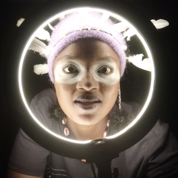 A woman with dark skin is centered within a bright, circular light. She wears a lavender knit cap adorned with white feathers, possibly a headdress. White paint or makeup is applied around her eyes. She also wears a necklace with pink and black beads and fluffy, dark earrings. Her expression is intense. The background is stark black, emphasizing the illuminated circle and the woman within.