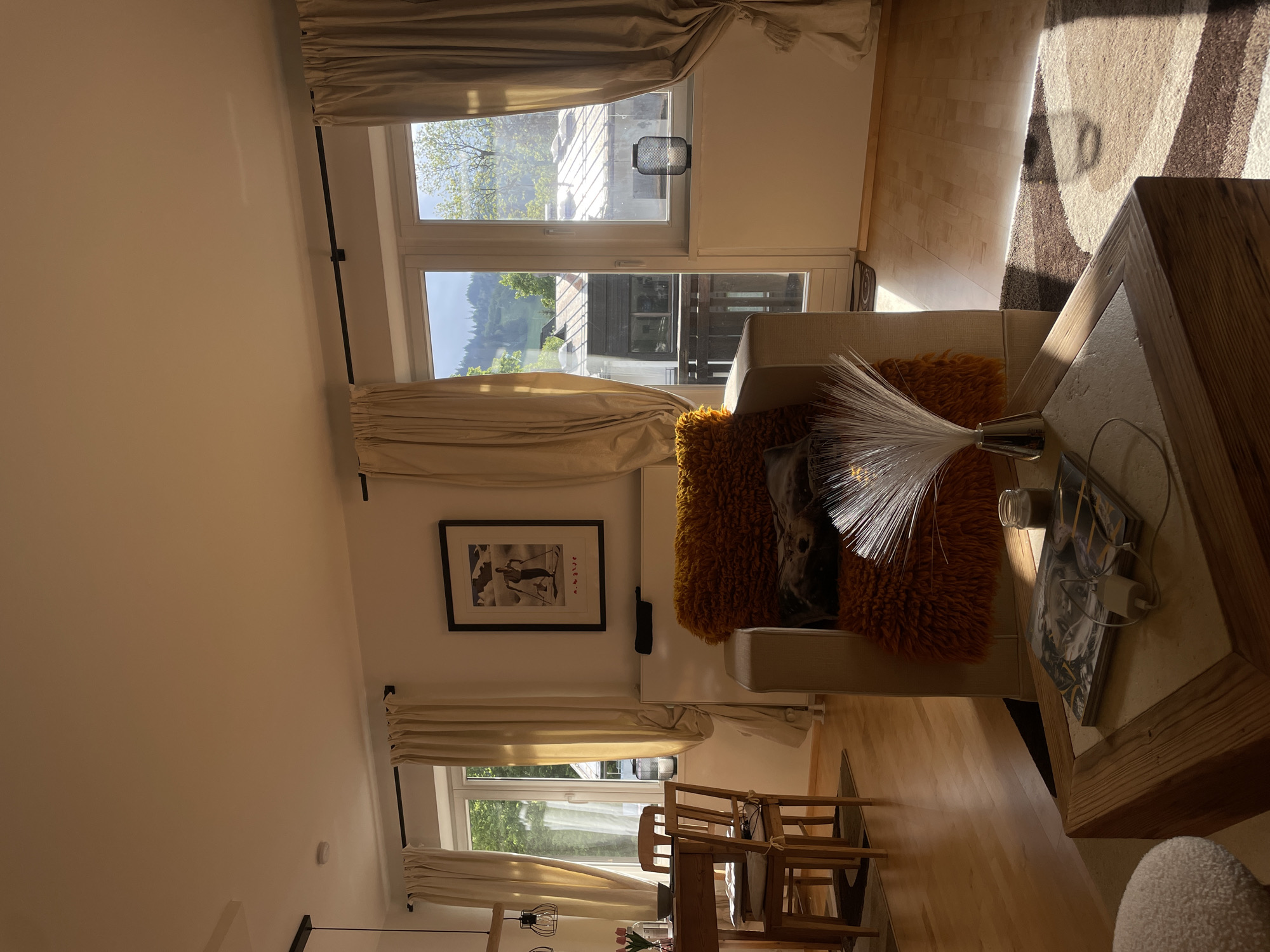 Here is a description of the image:
The image shows an interior living space in an unknown location, likely a home. Natural light floods the room through large windows dressed with neutral-toned curtains. There is a chair with a decorative orange pillow and a fiber-optic lamp on a wooden-framed table. Artwork hangs on the wall near the chair. A dining table and chairs are visible in the background. The floor is made of light-colored wood.